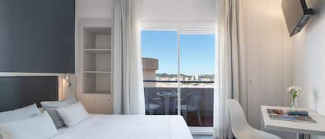 A hotel room with a bed, a desk, and a view of the city through large windows.