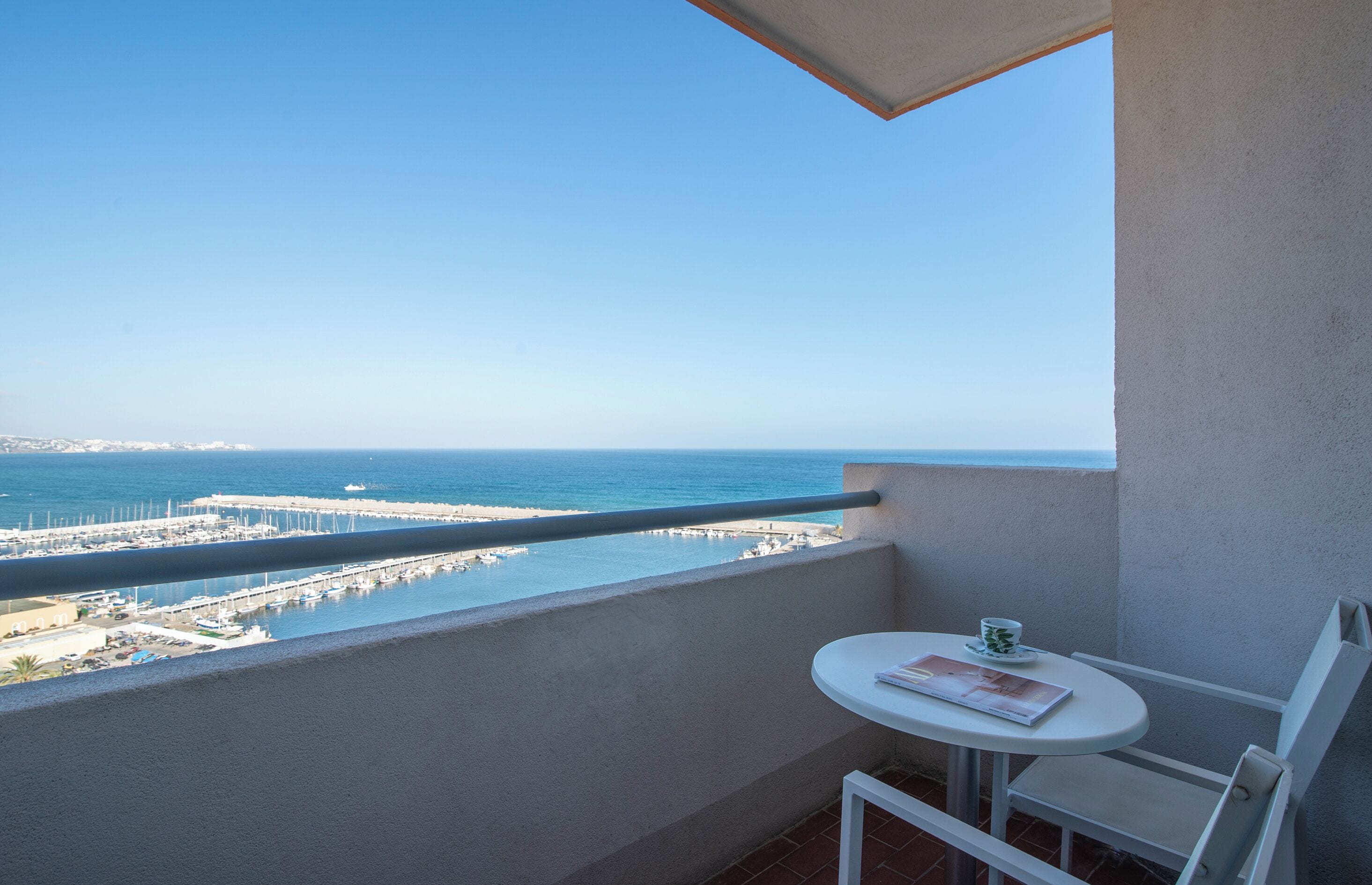 A balcony with a view of the sea, a small table with a chair, and a railing.