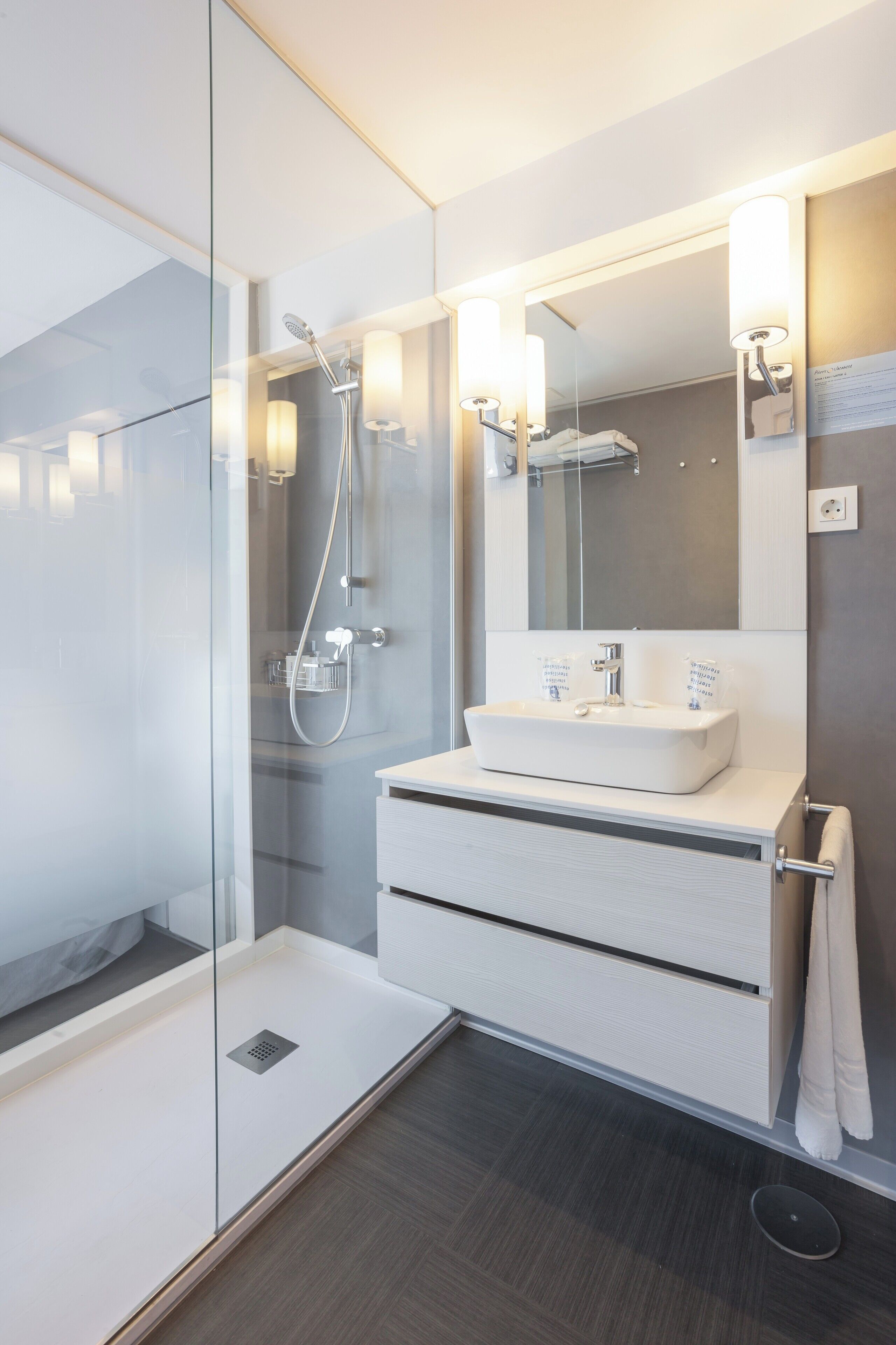 A modern bathroom with a glass shower enclosure, a white vanity with a large mirror, and a dark wood floor.