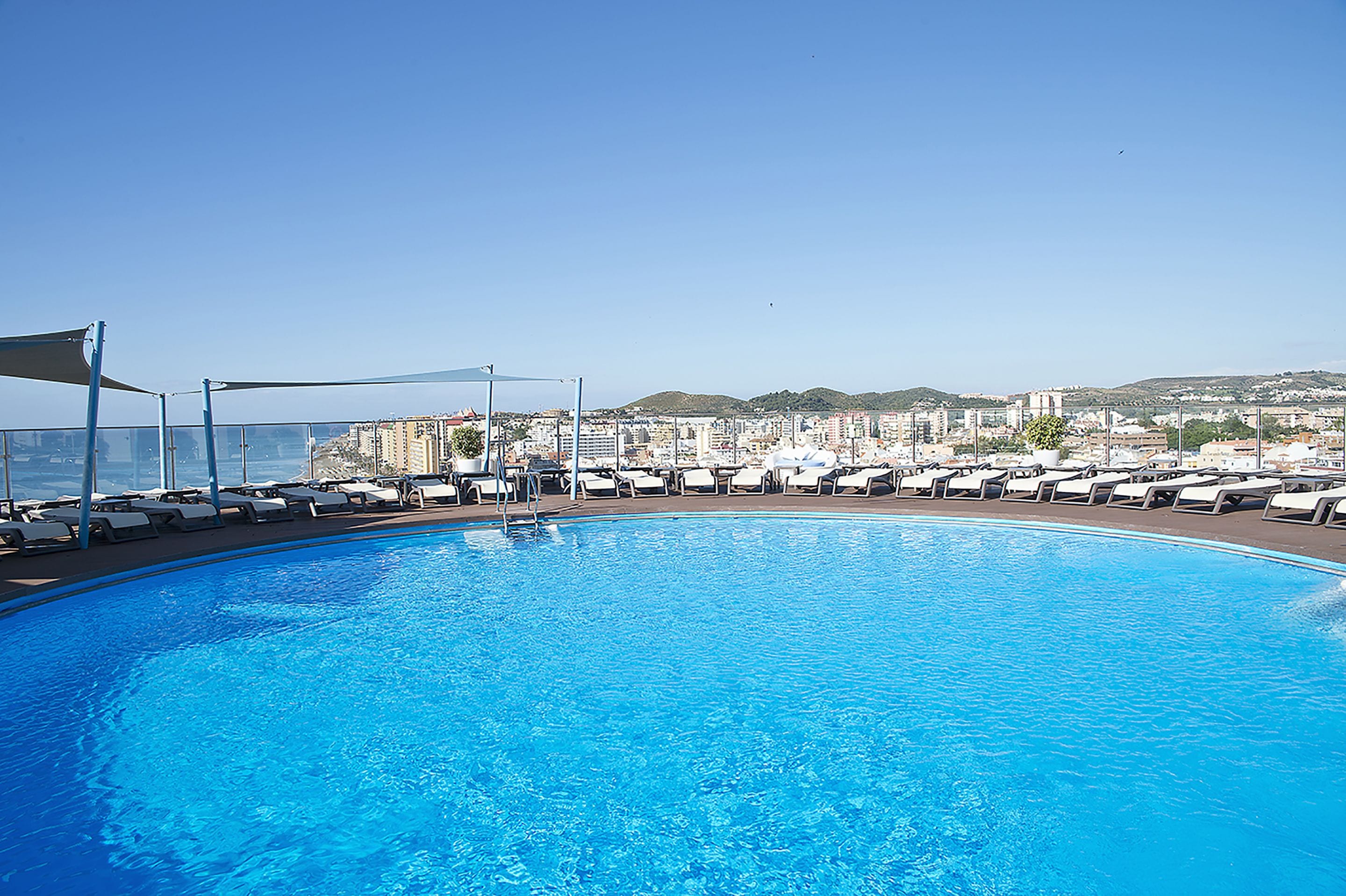 Una amplia piscina al aire libre con sillones de descanso y vistas a un paisaje urbano de fondo.
