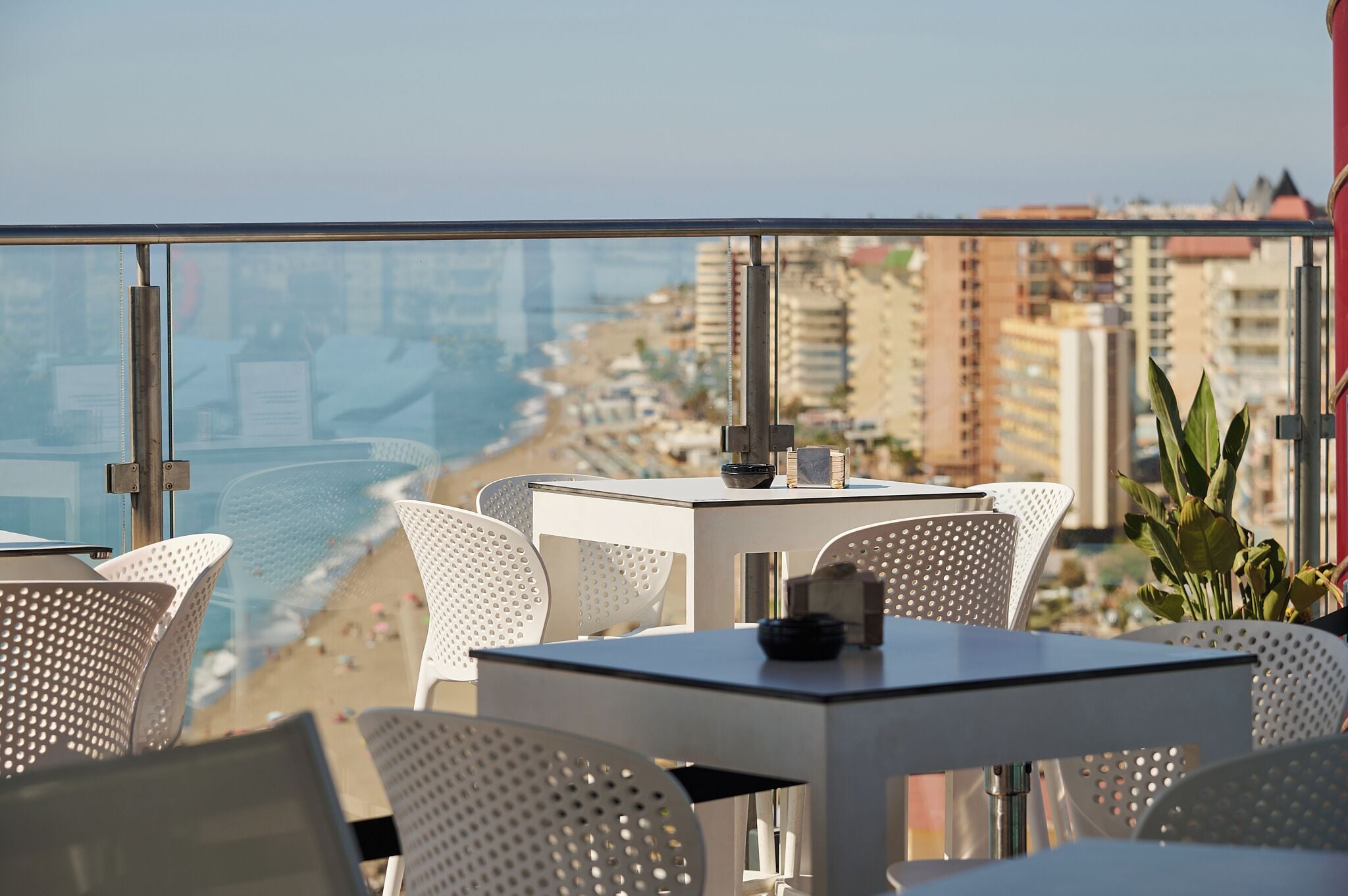 Un balcón con vistas a la playa y al paisaje urbano, con sillas y mesas blancas con diseño perforado.