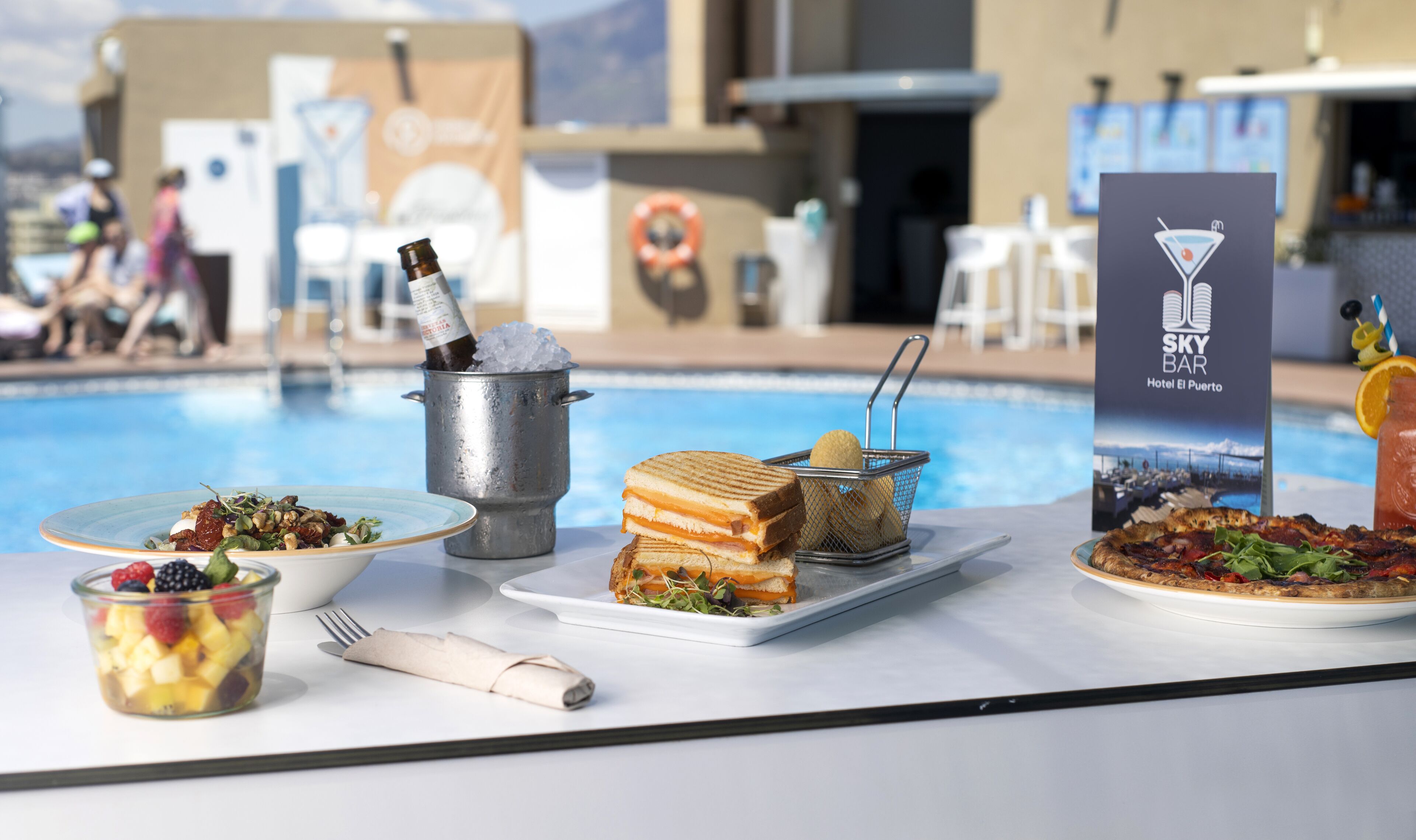 Disposición para comer junto a la piscina con diversos alimentos y bebidas, incluyendo un sándwich, un cuenco de frutas y un martini.