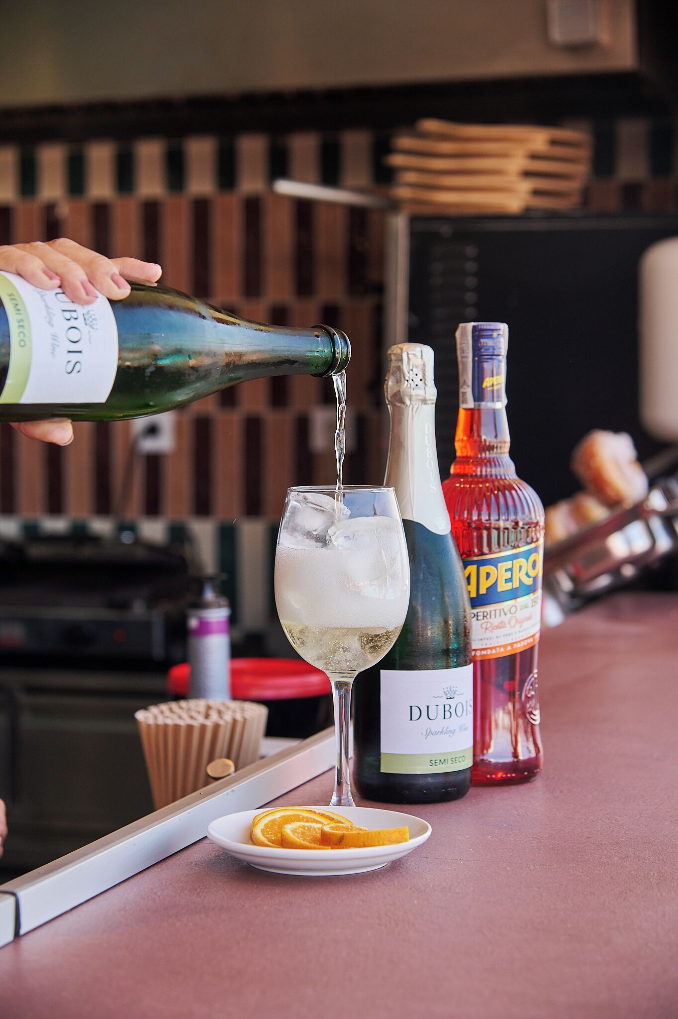 Une bouteille dont on verse le contenu dans un verre, une bouteille de Dubos posée sur le comptoir et une assiette avec des tranches d’orange.