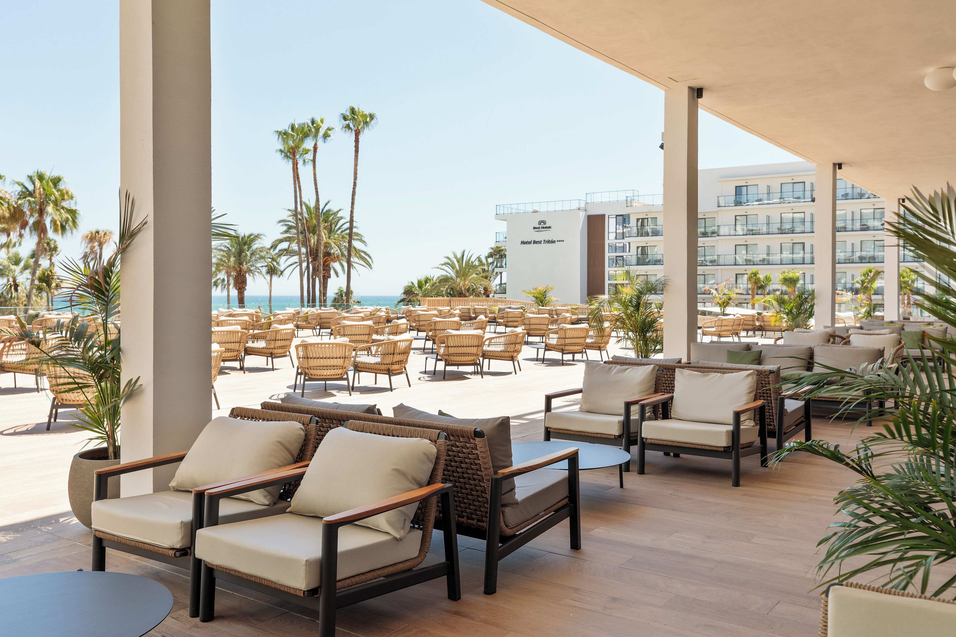 Une terrasse d’hôtel meublée de mobilier en osier, avec une piscine et une vue sur l’océan et les palmiers.