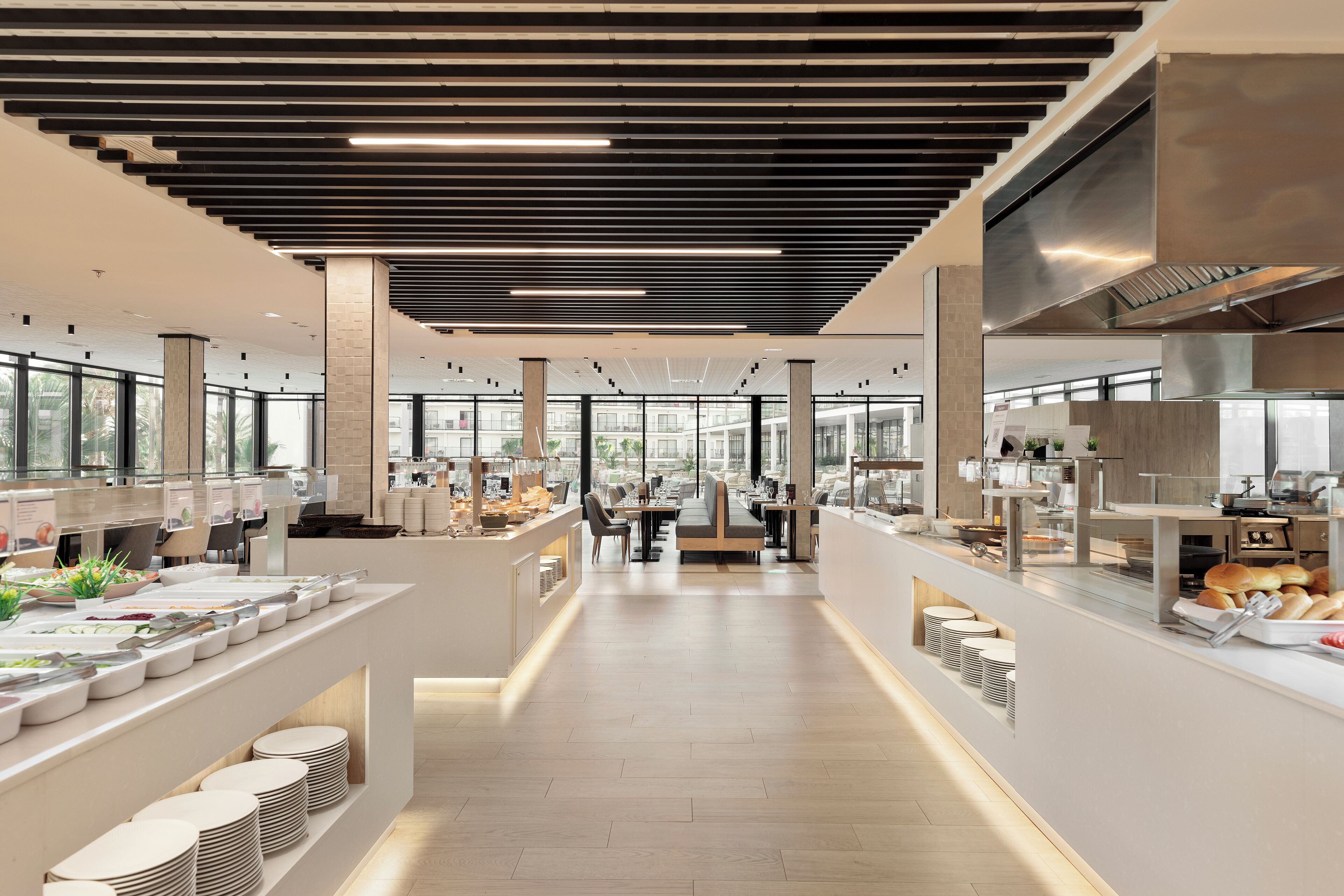 Una cafeteria moderna amb taules comunals llargues, una zona central de seients i una cuina al fons.