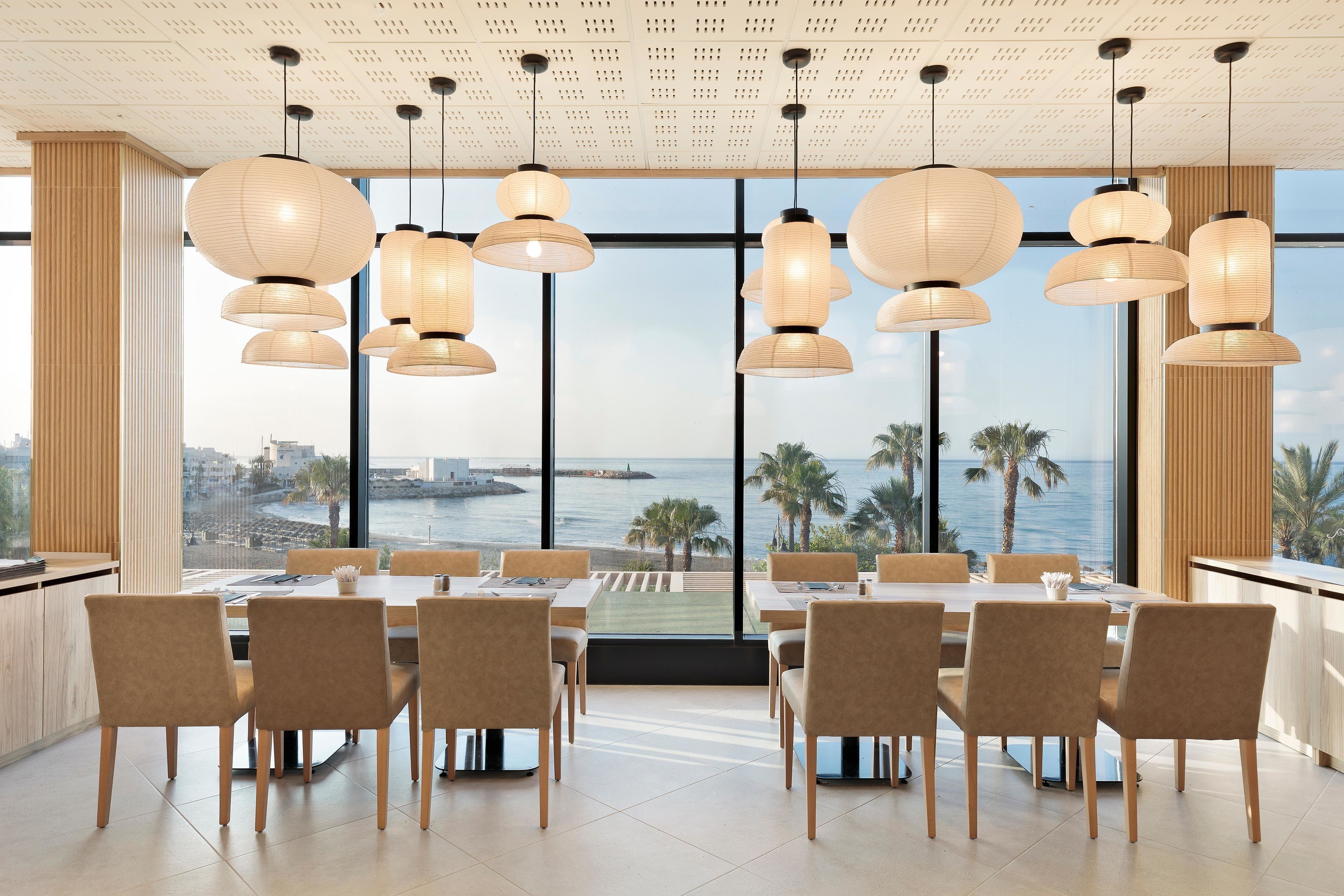 A modern dining area with large windows, multiple hanging light fixtures, and a view of the sea.