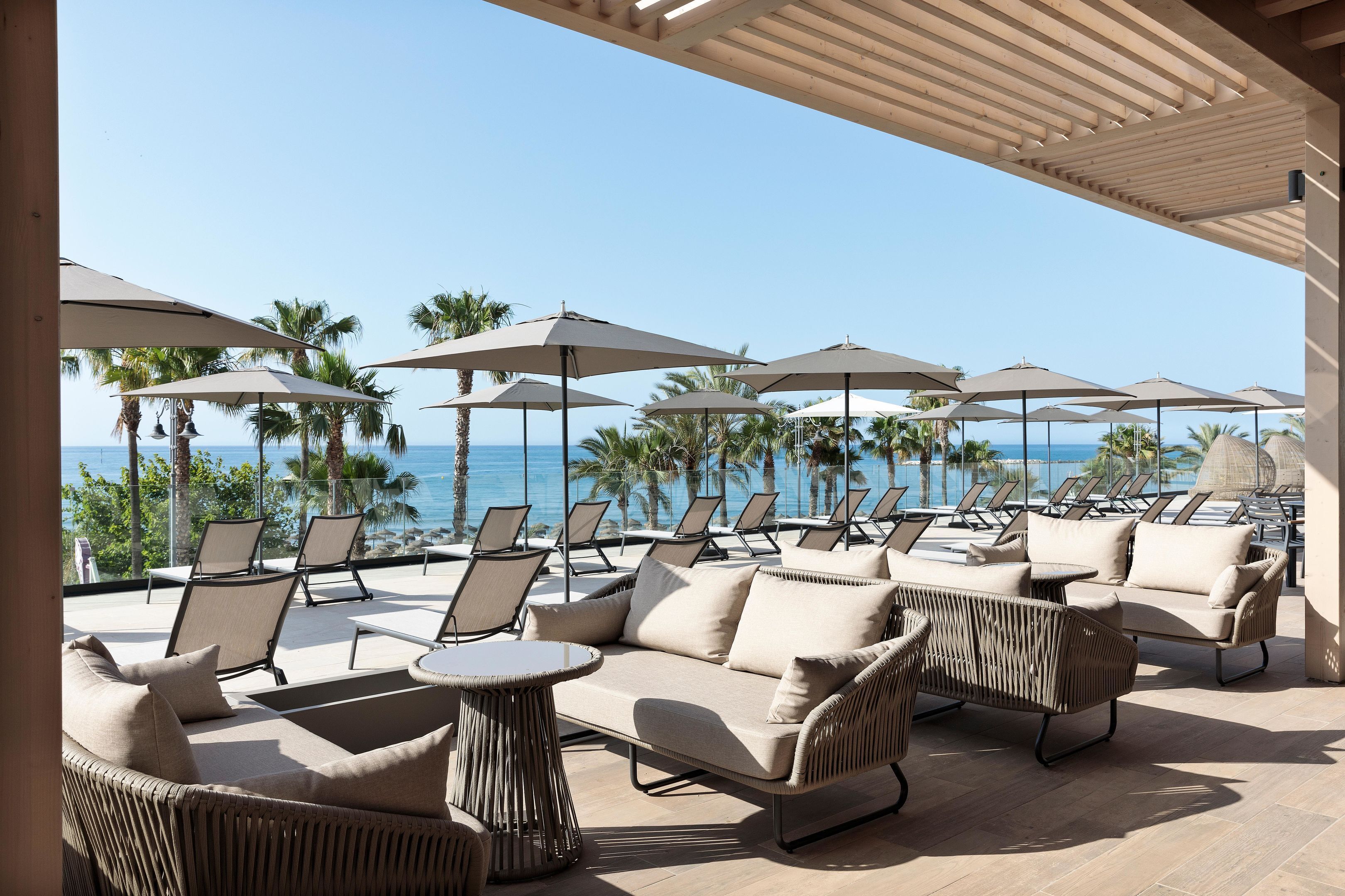 Un área de estar al aire libre con sillones reclinables y sombrillas, con vista a una piscina y palmeras.