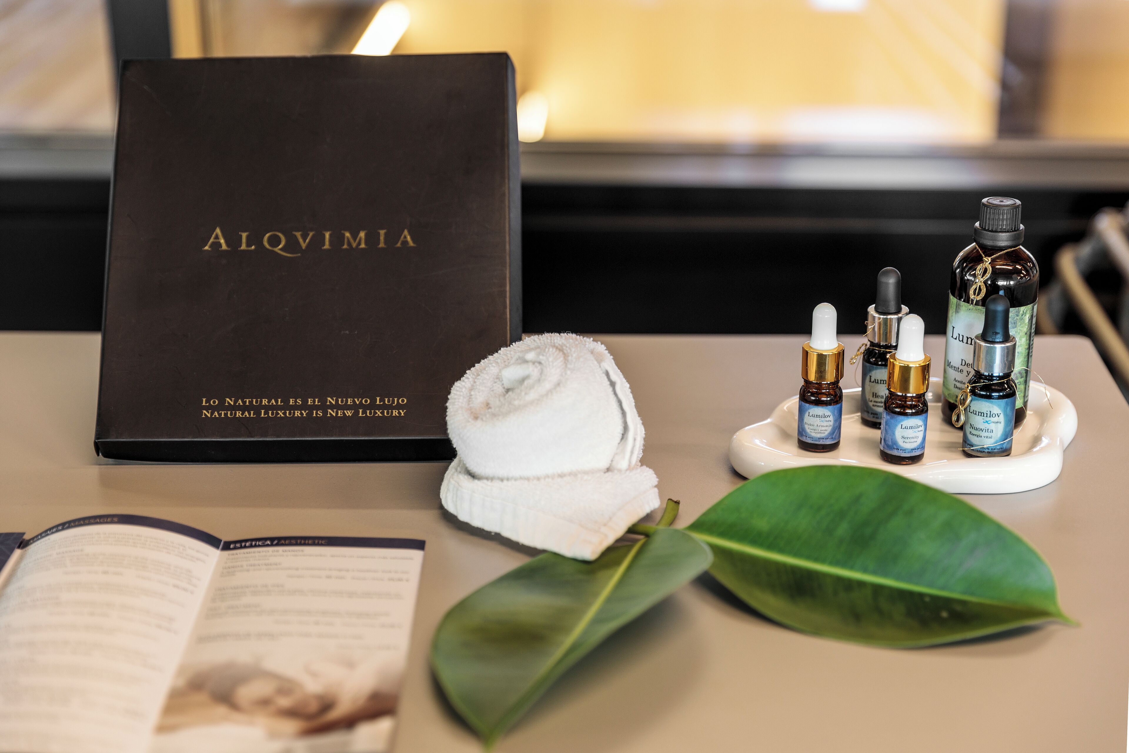 A luxury spa setting with a book titled 'ALQVIMIA', a rolled towel, and several bottles of 'Lumilov' skincare products on a tray.