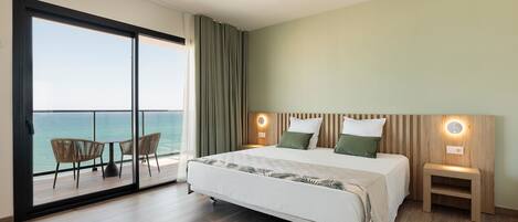 A hotel room with a bed, a nightstand, a small table with chairs, and a view of the ocean through a sliding glass door.