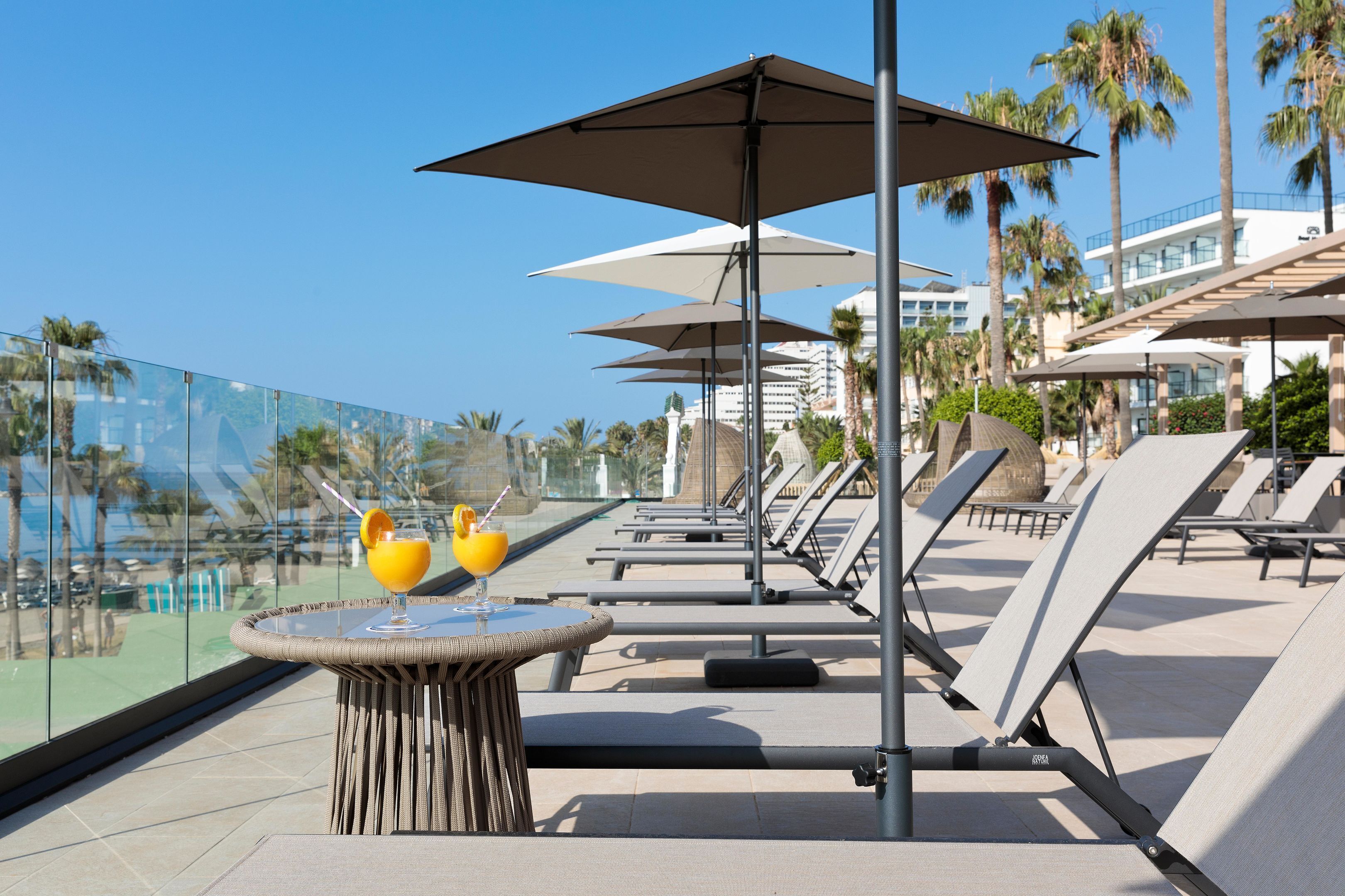 A rooftop pool area with lounge chairs and umbrellas, a table with drinks, and a clear view of the ocean and palm trees.