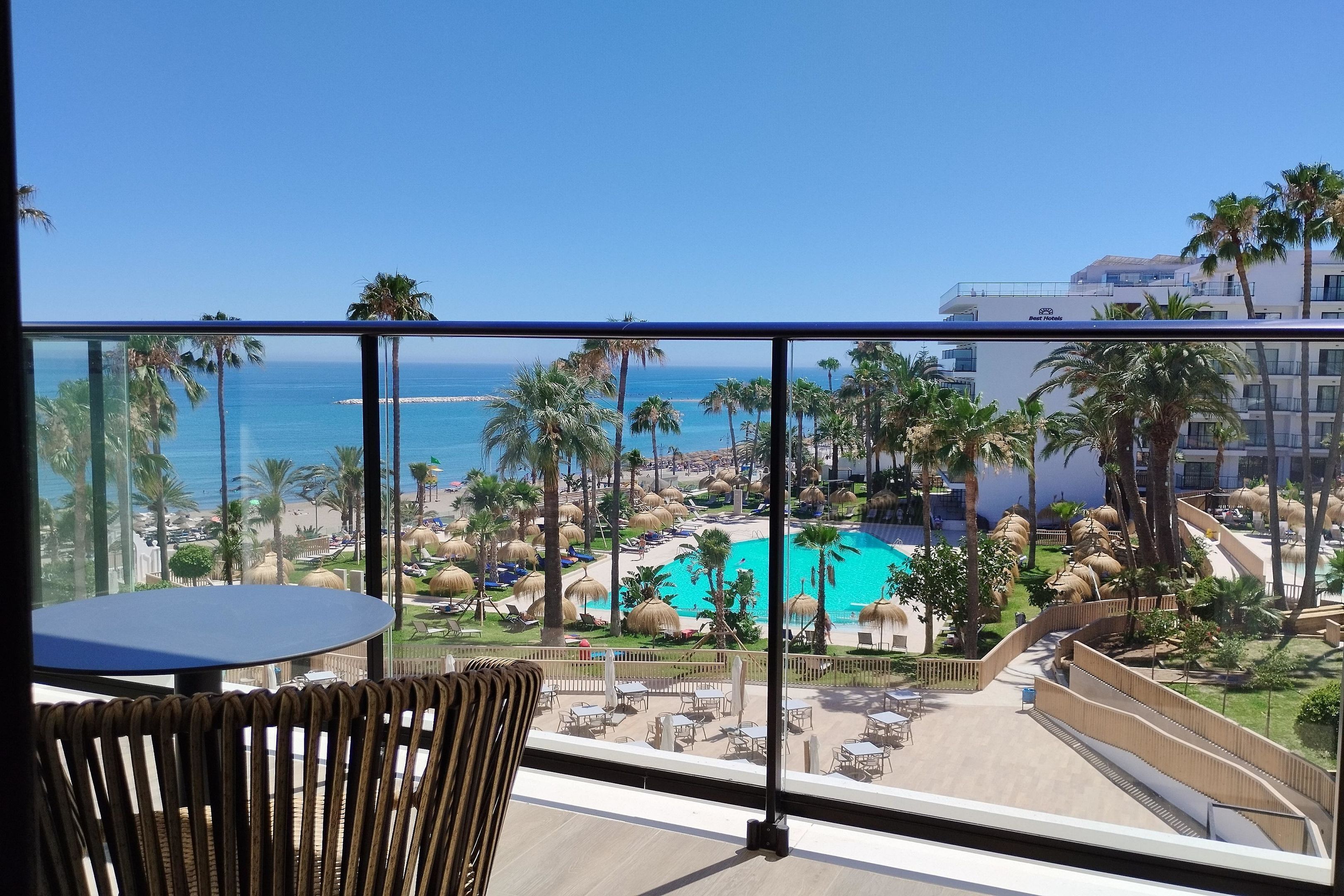A balcony with a view of a resort pool, palm trees, and a beach.