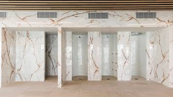 Un baño amplio y moderno con paredes y suelo de mármol, que cuenta con una zona de ducha con mampara de vidrio y un cabezal de ducha visible.