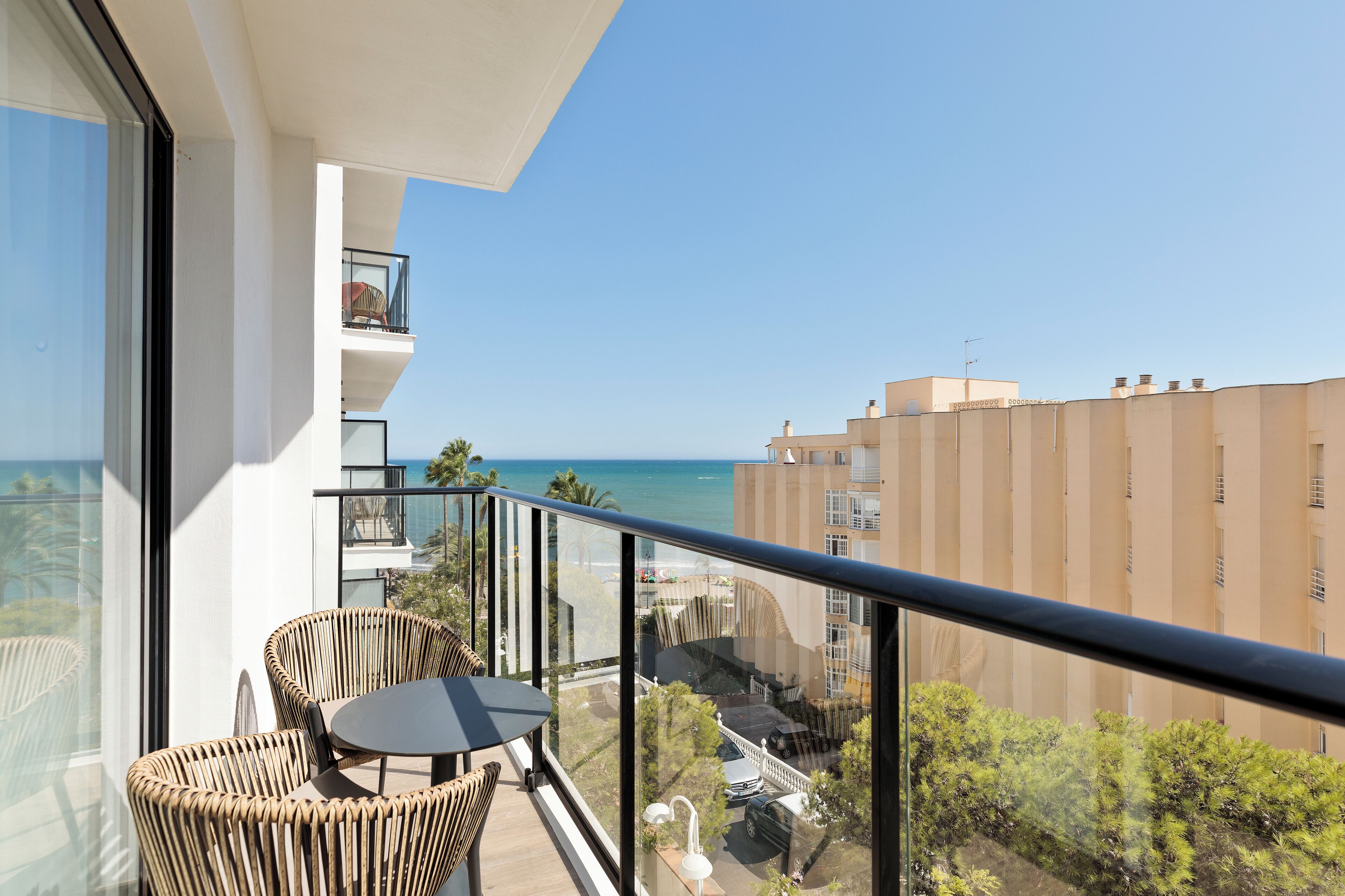Un balcón con sillas de mimbre y una mesa, con vistas a la costa, edificios y vegetación.