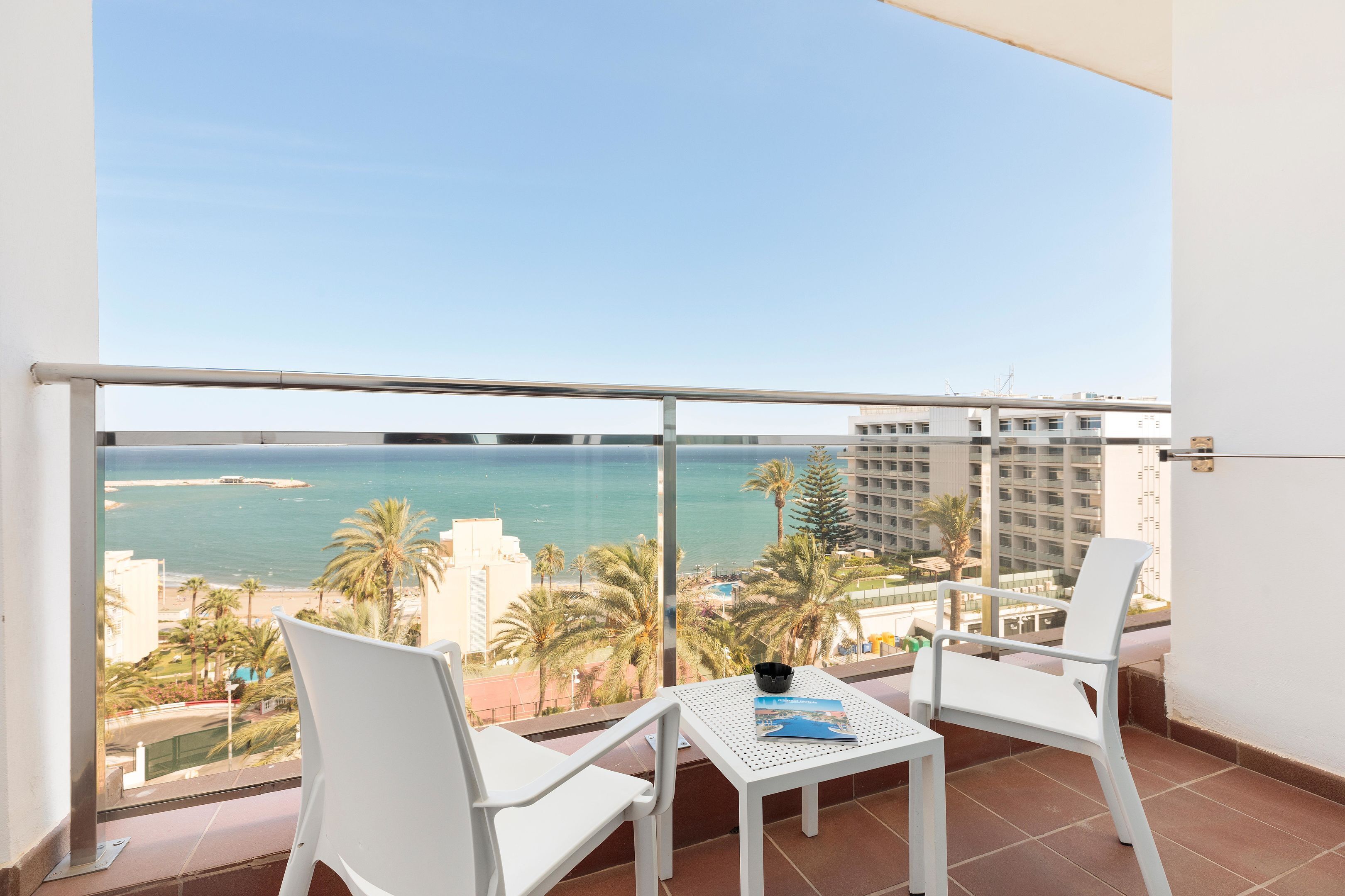 Un balcón con dos sillas blancas y una mesa, con vista a una playa y al océano, con palmeras y edificios al fondo.