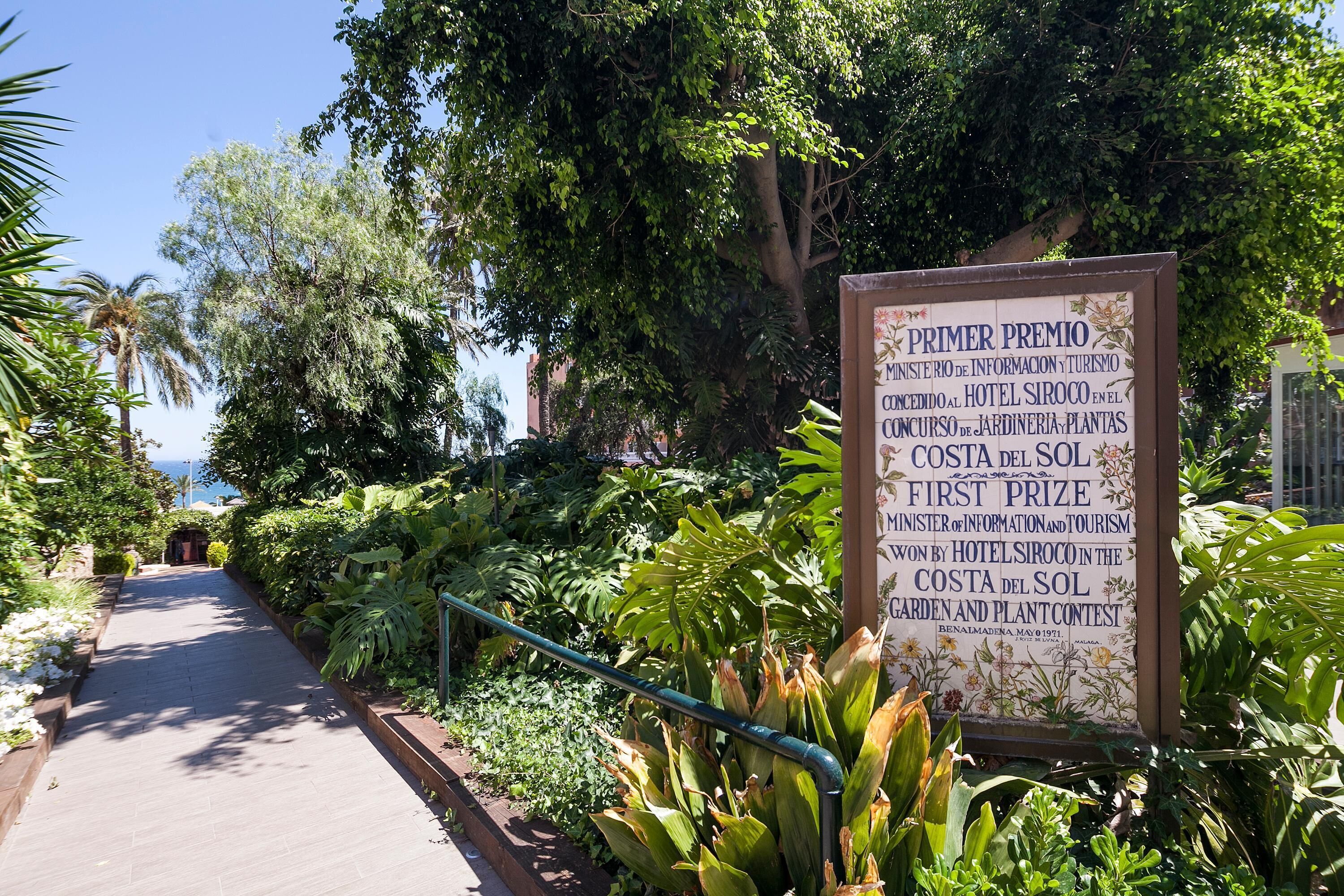 Un letrero con información sobre un concurso de jardines y plantas, rodeado de abundante vegetación y un sendero.