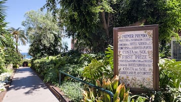 Un letrero con información sobre un concurso de jardines y plantas, rodeado de abundante vegetación y un sendero.