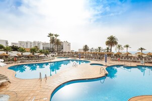 En swimmingpool med liggestole og palmer, omgivet af bygninger.