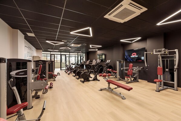 Un gimnasio con diversos equipos de ejercicio, incluyendo cintas de correr, máquinas de pesas y una pantalla de televisión mostrando 'KISS FM'.