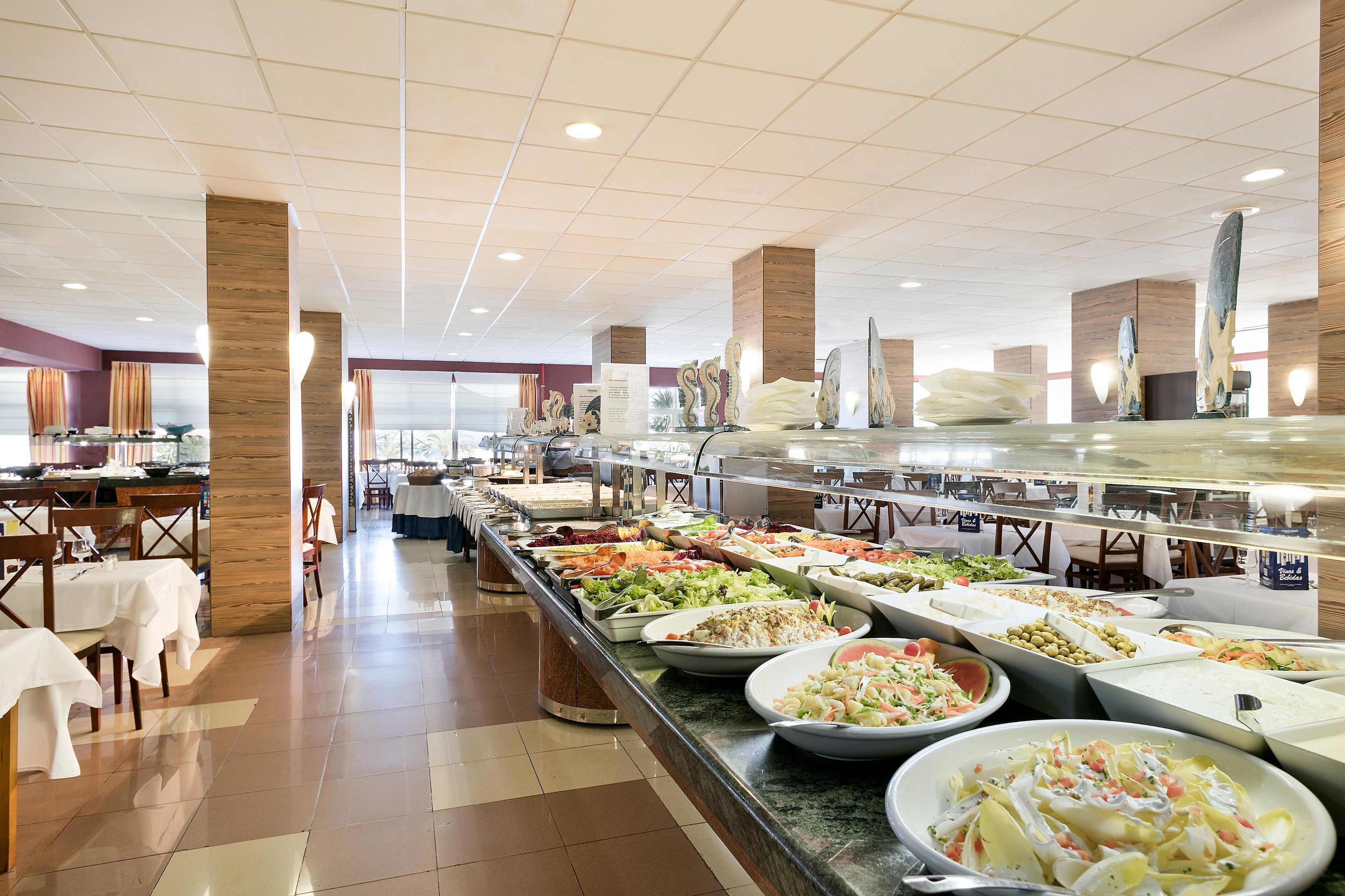 Buffet com diversas opções de comida, mesas e cadeiras dispostas numa área ampla com piso de azulejos e detalhes em madeira.