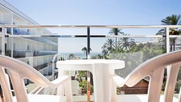 A balcony with white chairs and a table, overlooking a building with multiple balconies and palm trees.