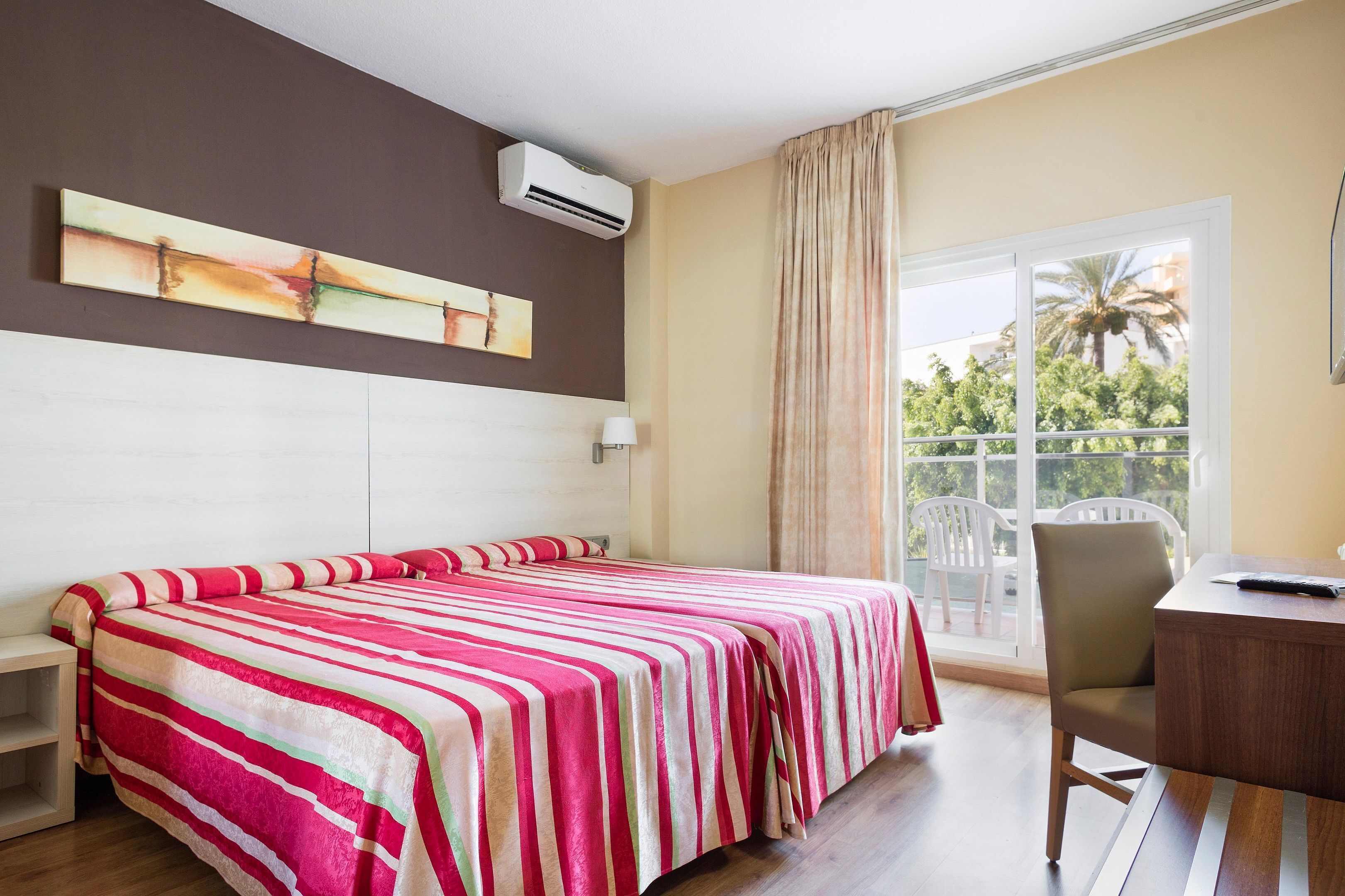 A hotel room with a bed, a desk, a chair, a window with a view of palm trees, and an air conditioning unit.