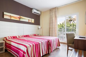 Une chambre d’hôtel équipée d’un lit, d’un bureau, d’une chaise, d’une fenêtre donnant sur des palmiers et d’un appareil de climatisation.