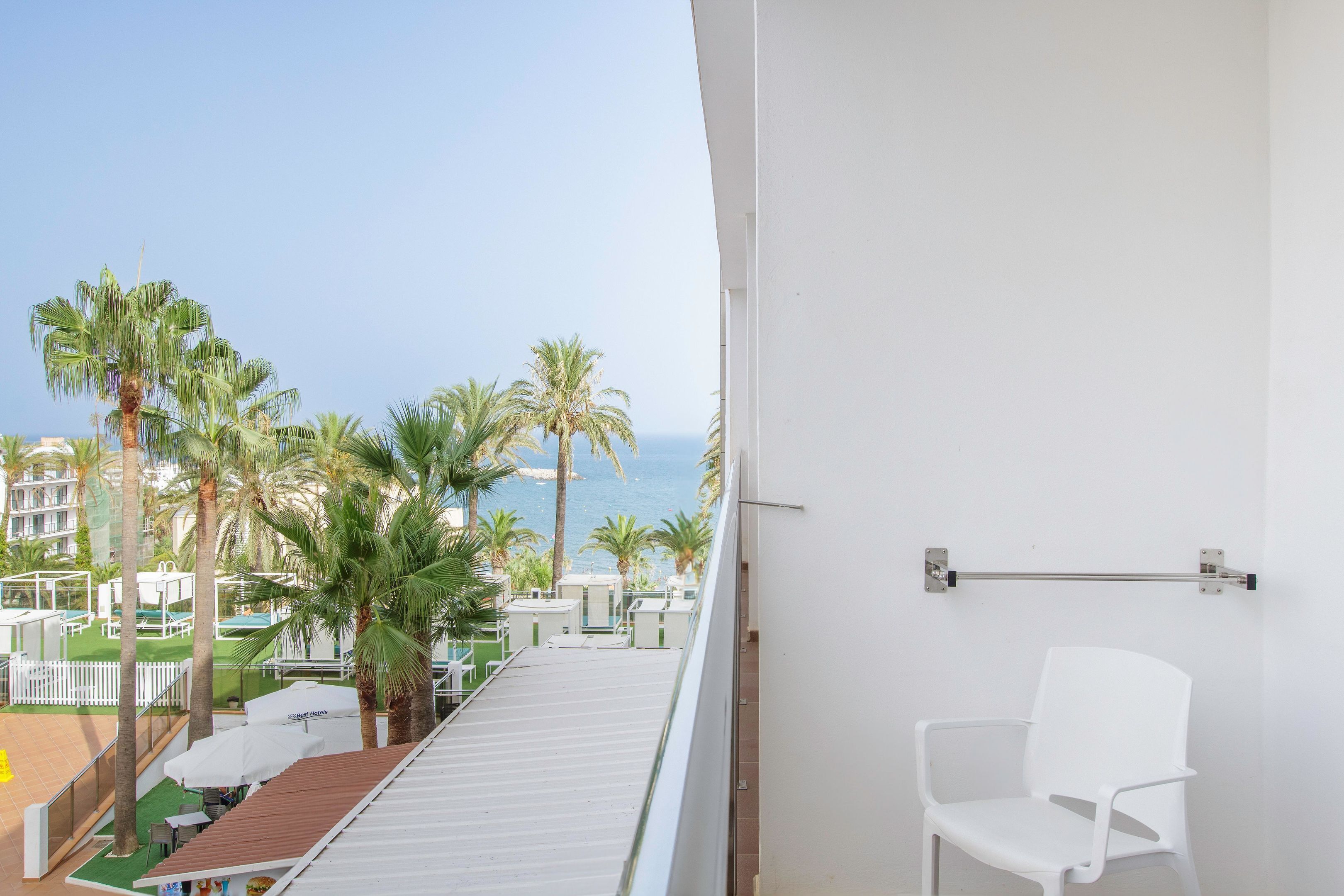 Een balkon met een witte stoel, uitkijkend op een zwembad met palmbomen en uitzicht op zee.