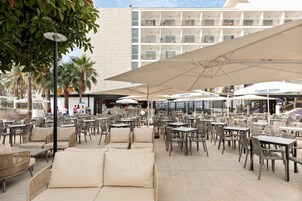 An outdoor cafe area with multiple tables and chairs, umbrellas, and a view of a hotel building in the background.