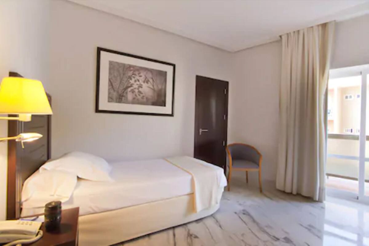 A hotel room with a bed, a desk with a lamp, a chair, a framed picture on the wall, and a door.