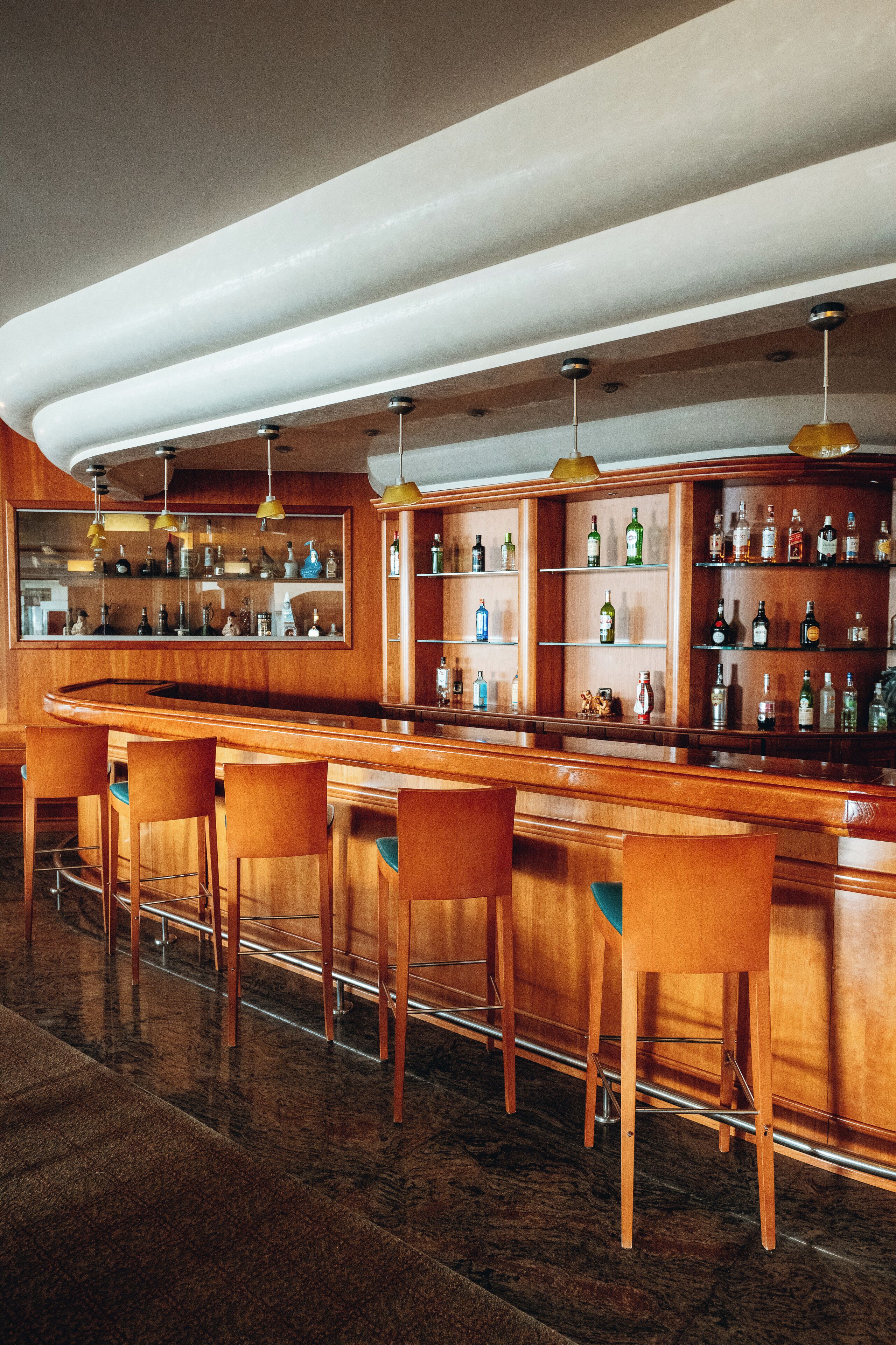 Zona de bar con mostrador de madera, varios taburetes y estantes con botellas.