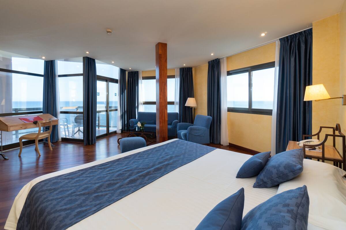 A hotel room with a large bed, a desk with a chair, a table with a chair, and a view of the sea through large windows.