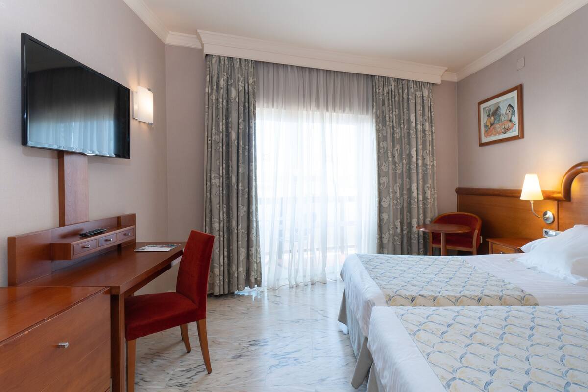 A hotel room with a bed, a desk with a chair, a television, a window with curtains, and a painting on the wall.