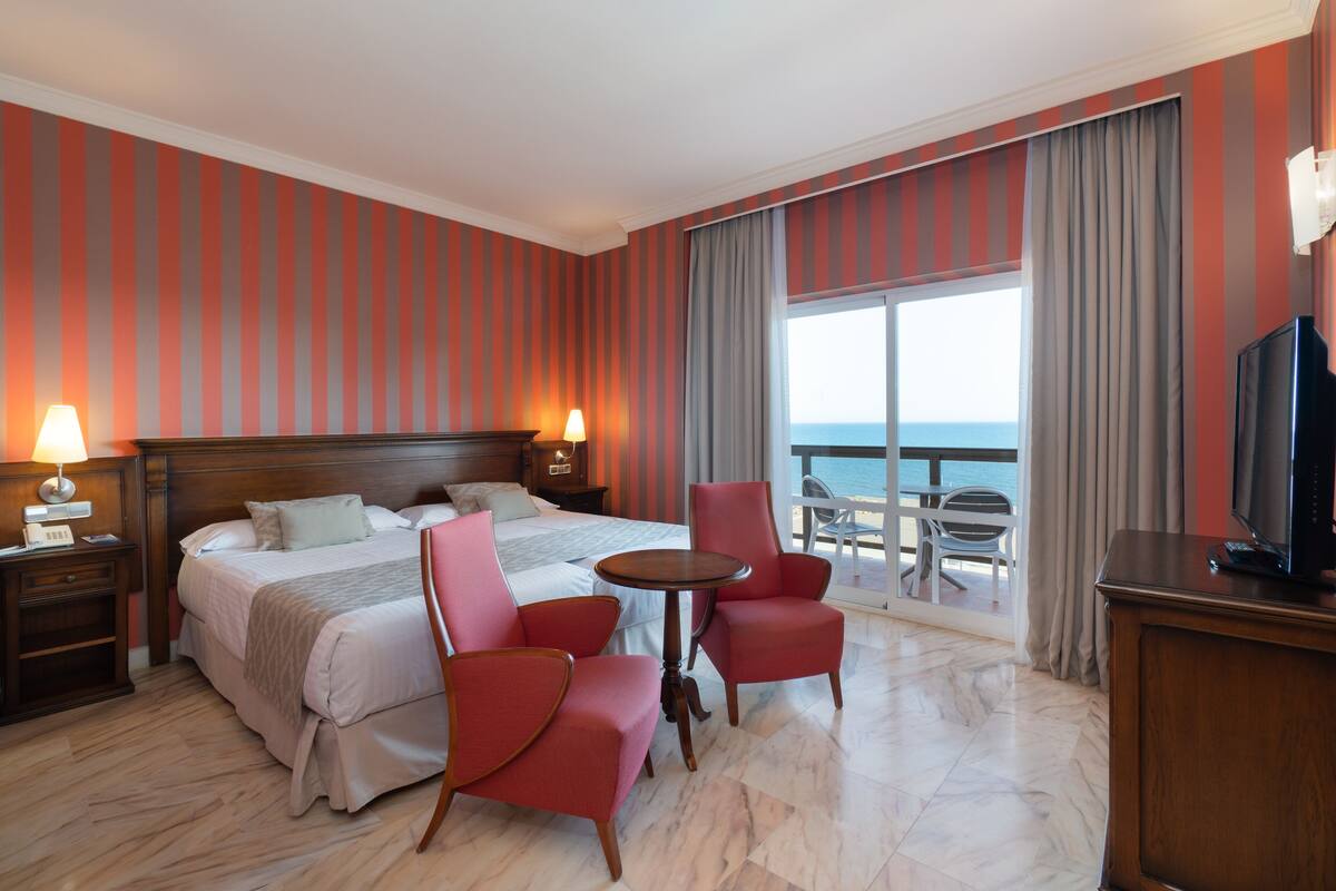 A hotel room with a bed, two red chairs, a small table, a TV, and a balcony with a view of the sea.