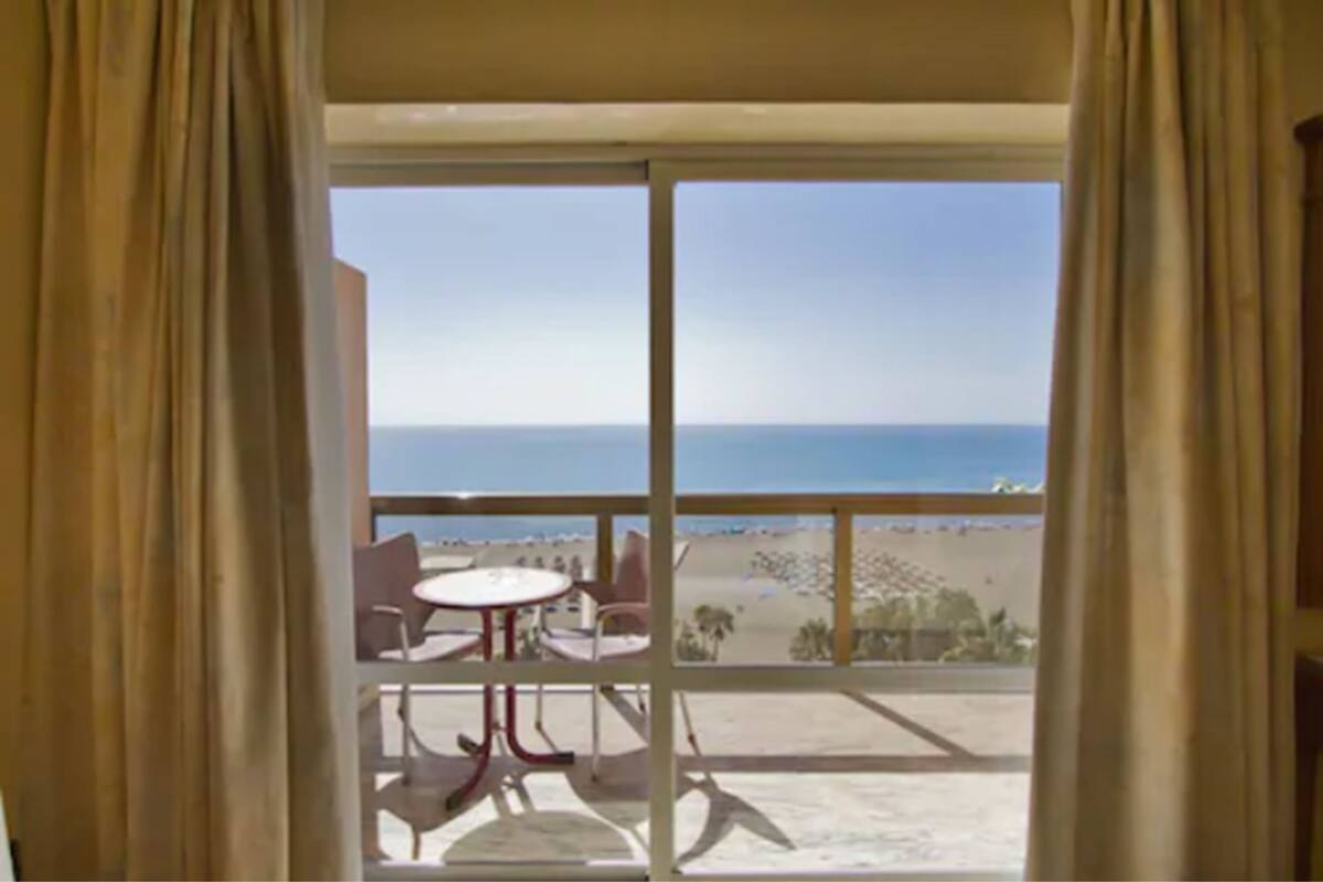 A balcony with a table and chairs, overlooking a beach and ocean.