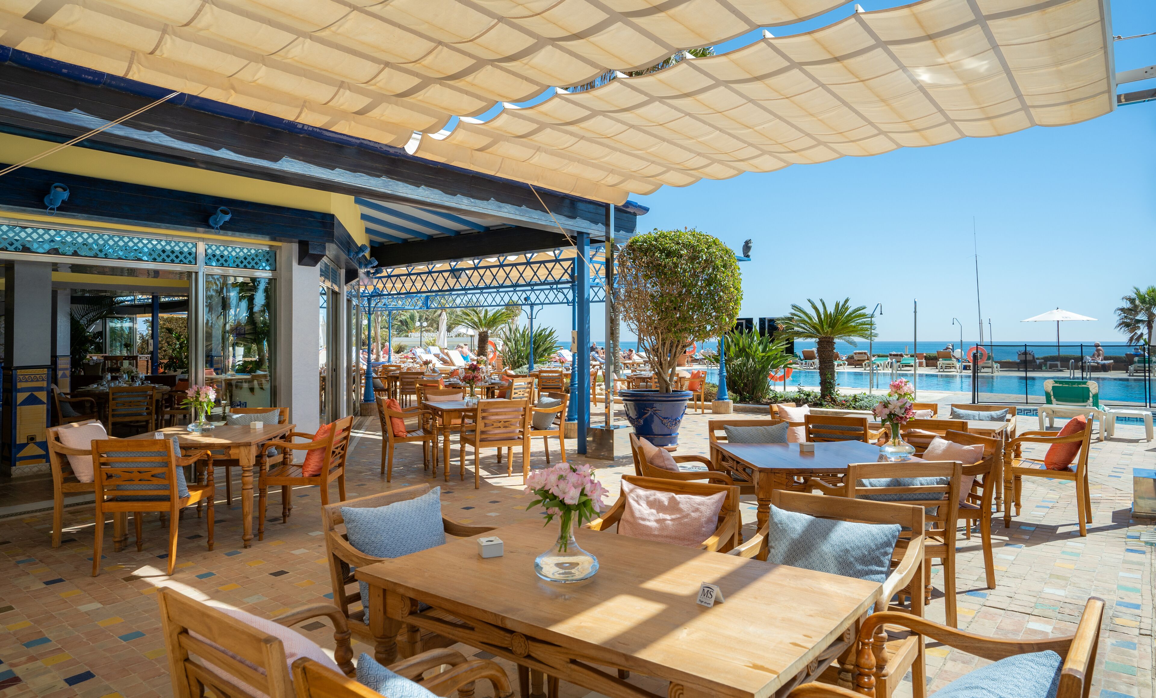 An outdoor seating area with wooden tables and chairs, a large potted plant, and a view of the pool and ocean.