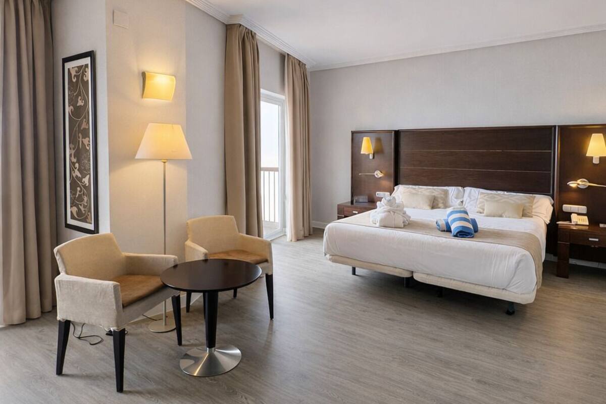 A hotel room with a bed, two chairs, a small table, and a lamp.