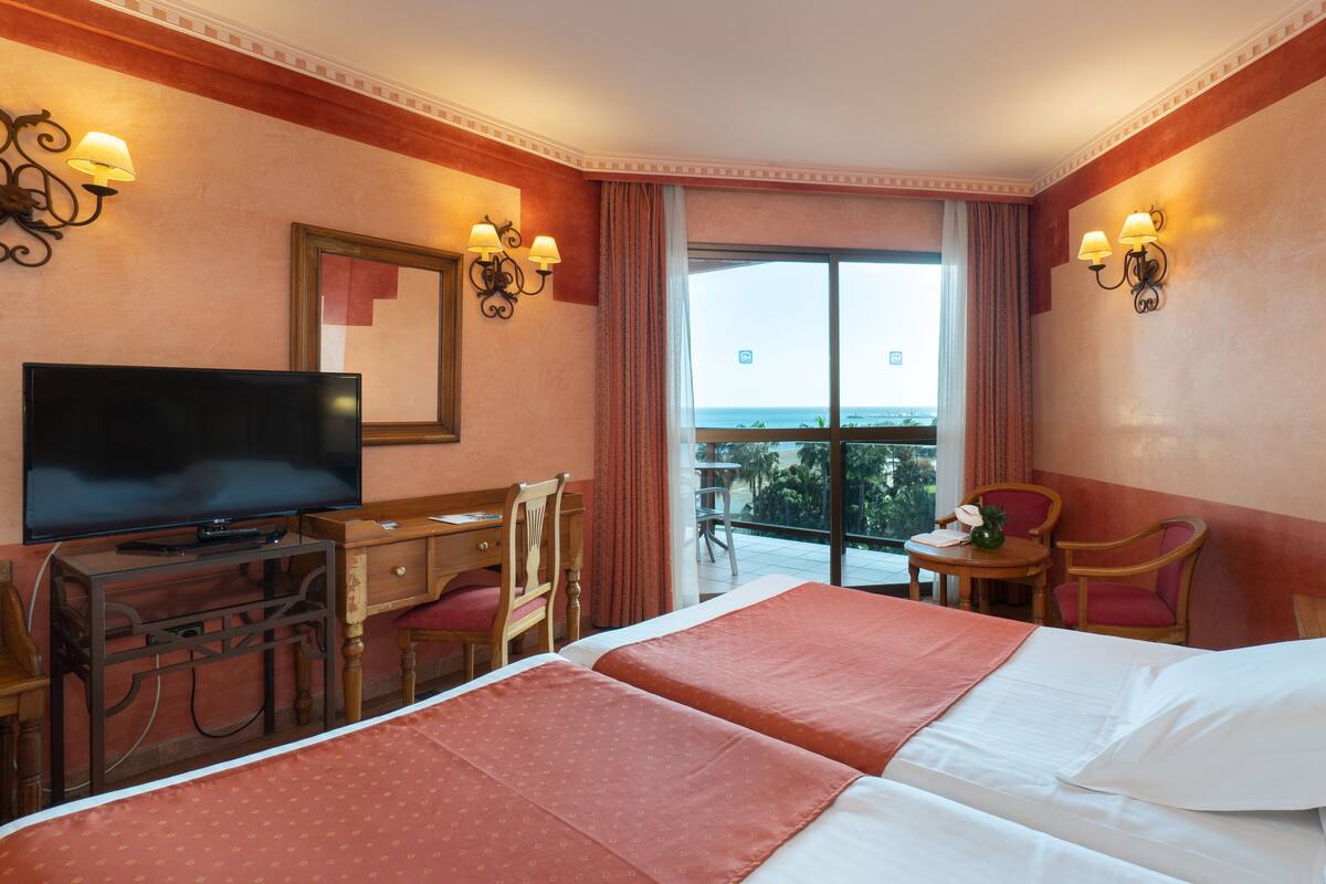 A hotel room with a bed, a desk with a TV, a chair, a small table with a lamp, and a view of the beach.