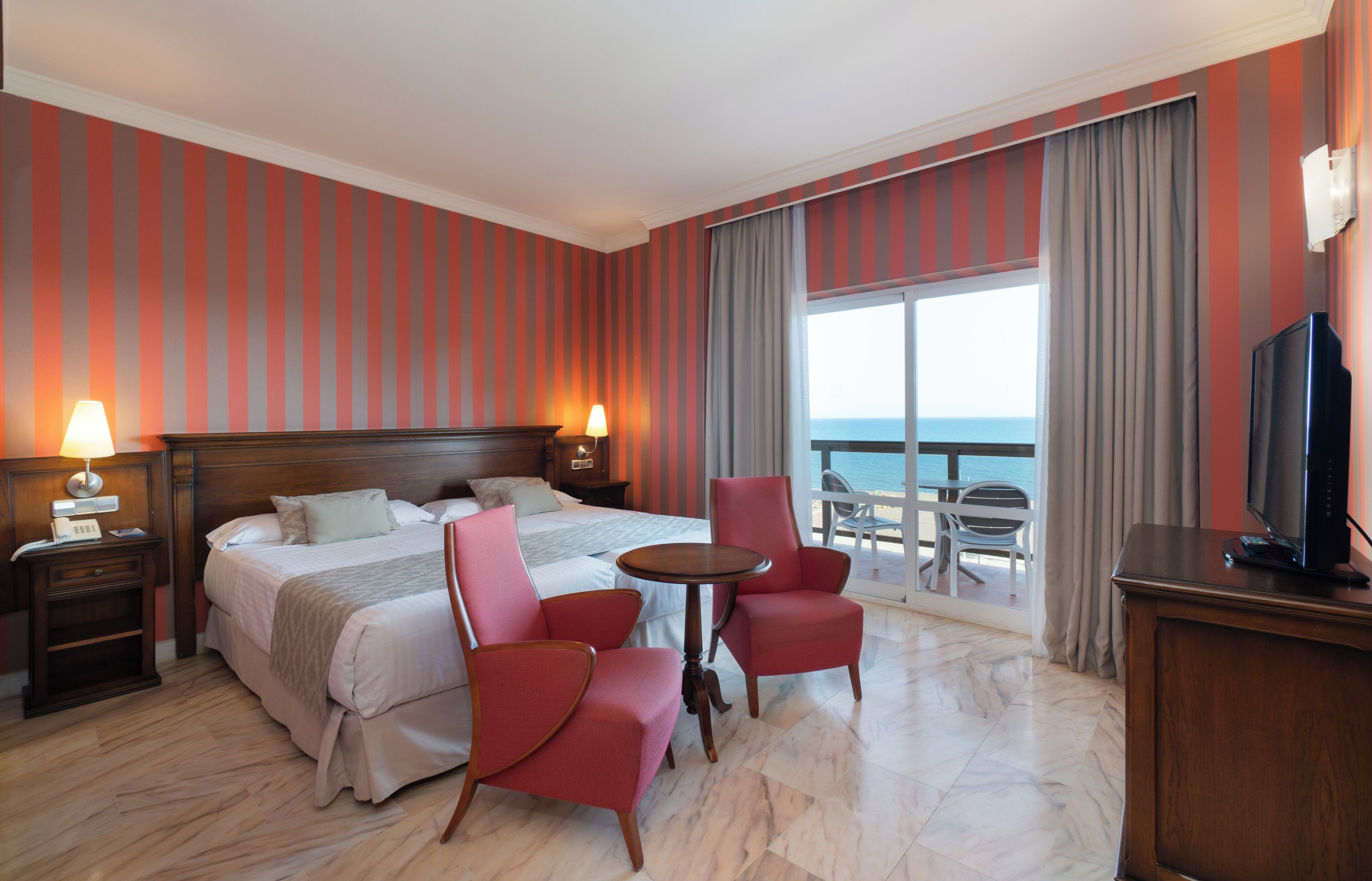 A hotel room with a bed, two red chairs, a small table, a TV, and a balcony with a view of the sea.