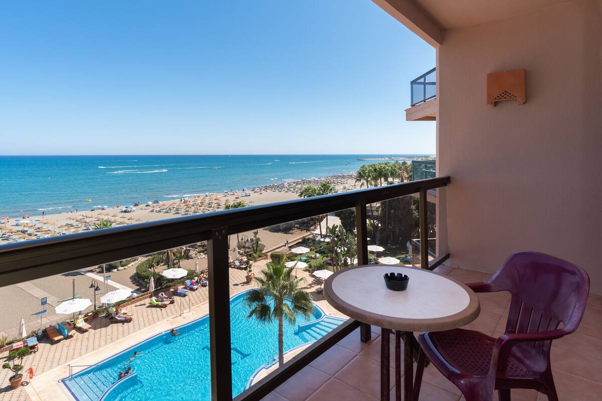A balcony with a pool, a table, and chairs overlooking a beach and ocean.