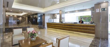A modern hotel lobby with a reception desk, seating area, and a staircase leading to an elevator.
