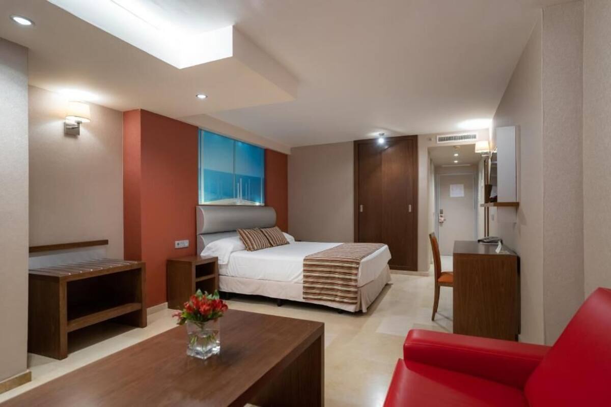 A modern hotel room with a bed, a desk, a red chair, and a small table with a vase of flowers.