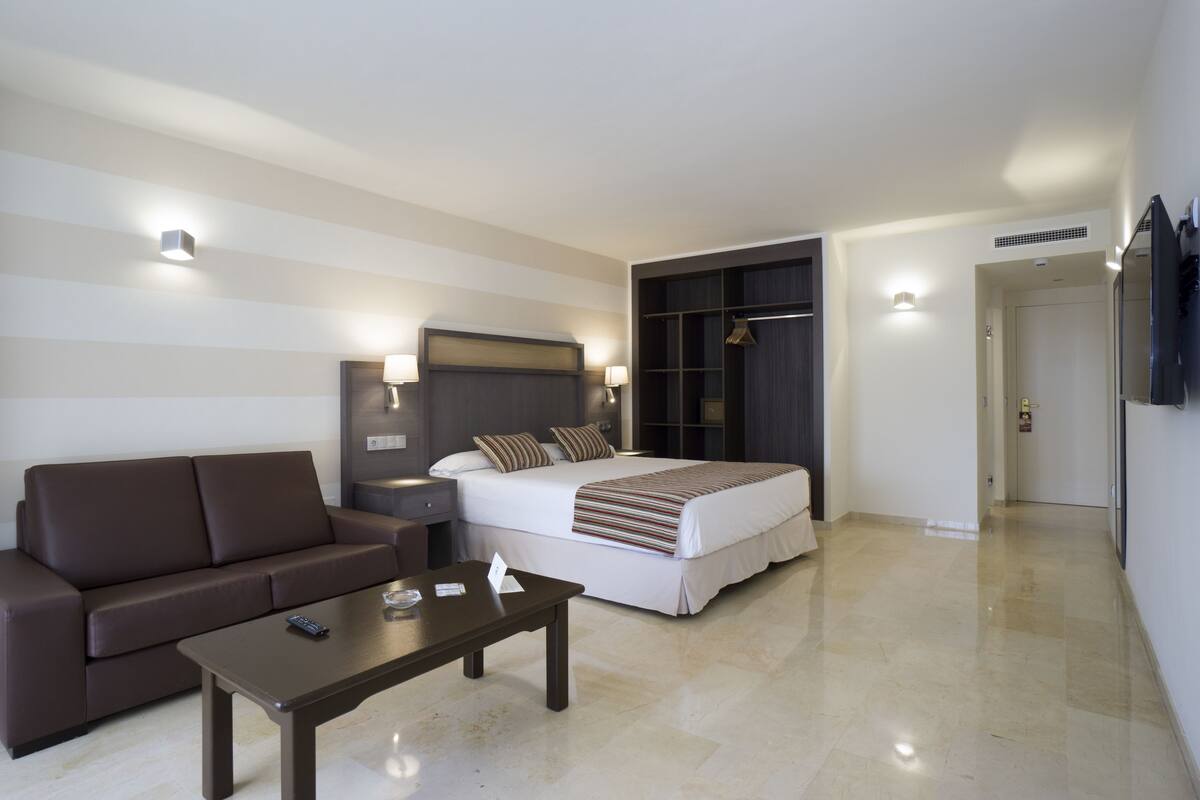 A hotel room with a sofa, a bed with a striped blanket, a bedside table, and a wardrobe.