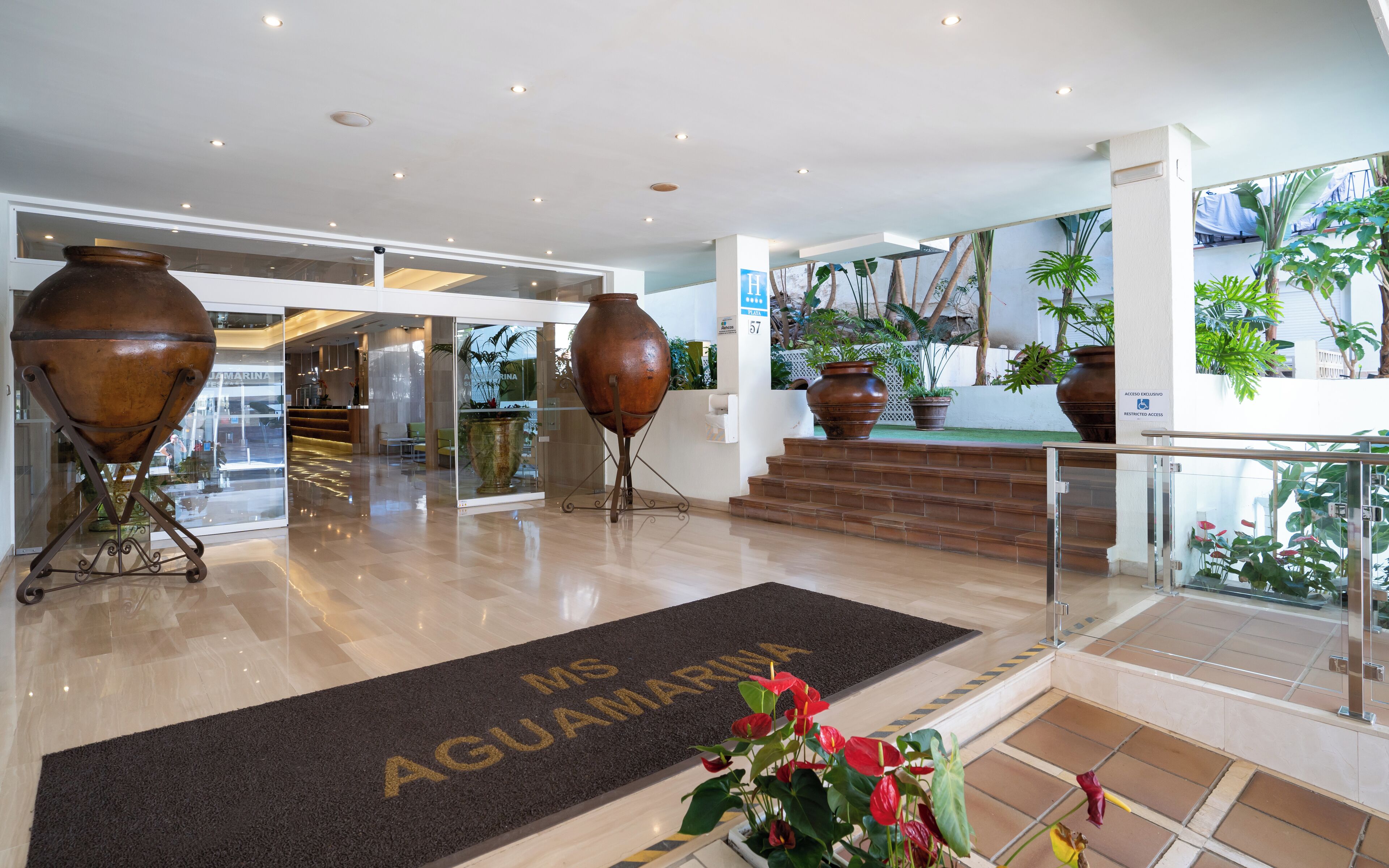 Een hotel lobby met een receptiebalie, grote vazen en een deurmat met de tekst 'MS AGUAMARINA'.