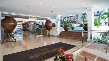 Een hotel lobby met een receptiebalie, grote vazen en een deurmat met de tekst 'MS AGUAMARINA'.