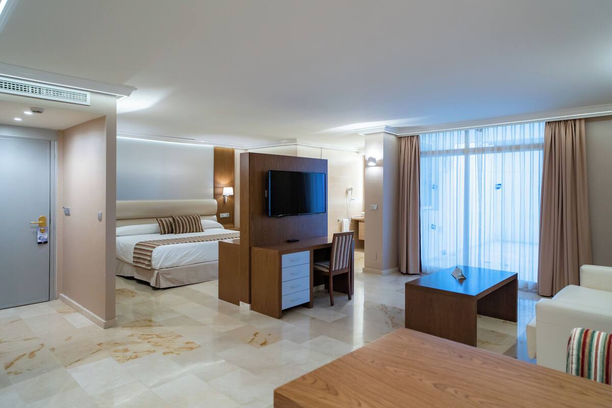 A hotel room with a bed, a TV, a desk, a chair, a sofa, and a large window with curtains.