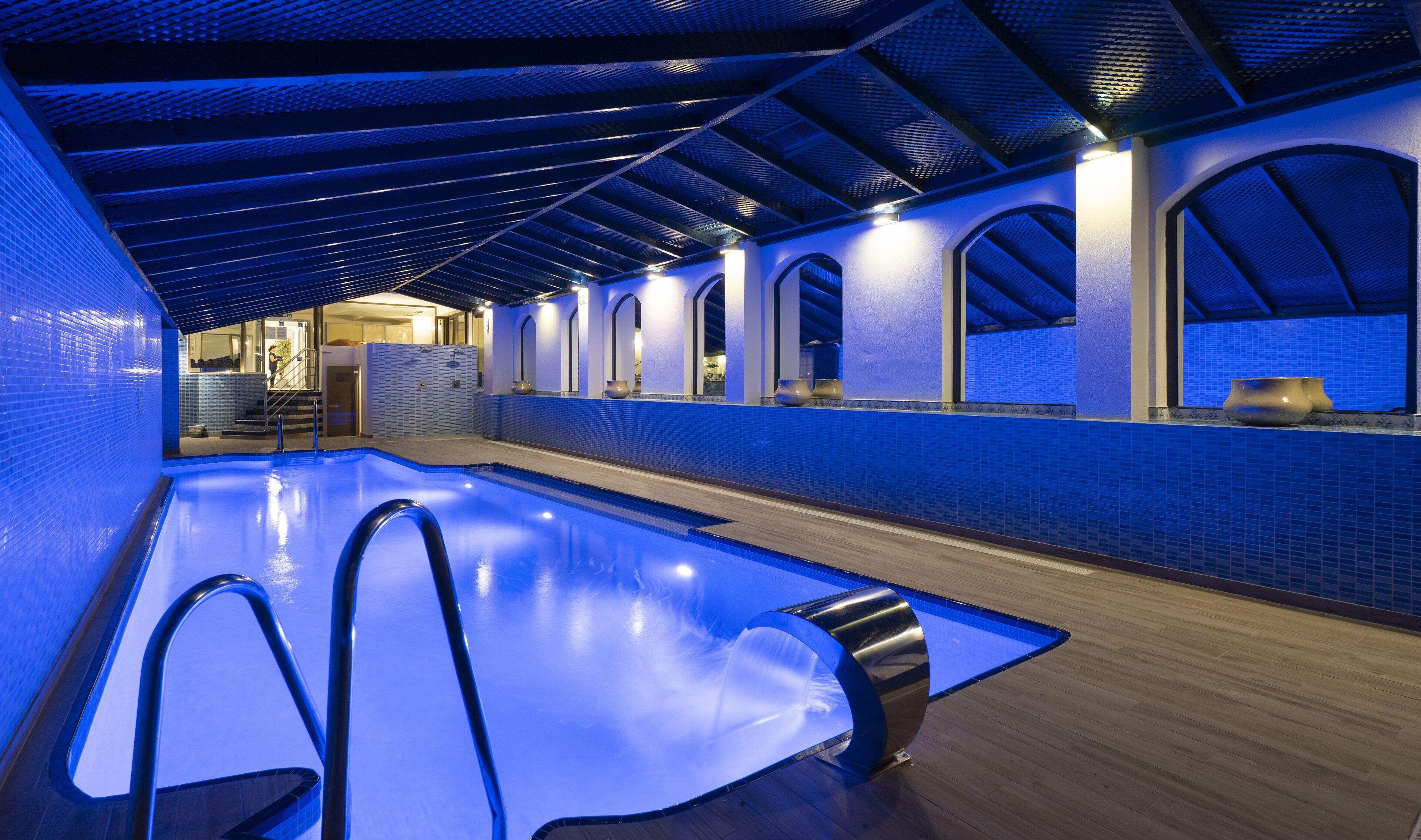 Une piscine intérieure moderne, dotée d’un éclairage bleu, de fenêtres en arc de cercle et d’un plancher en bois.