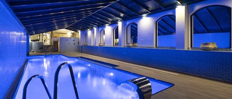 Une piscine intérieure moderne, dotée d’un éclairage bleu, de fenêtres en arc de cercle et d’un plancher en bois.