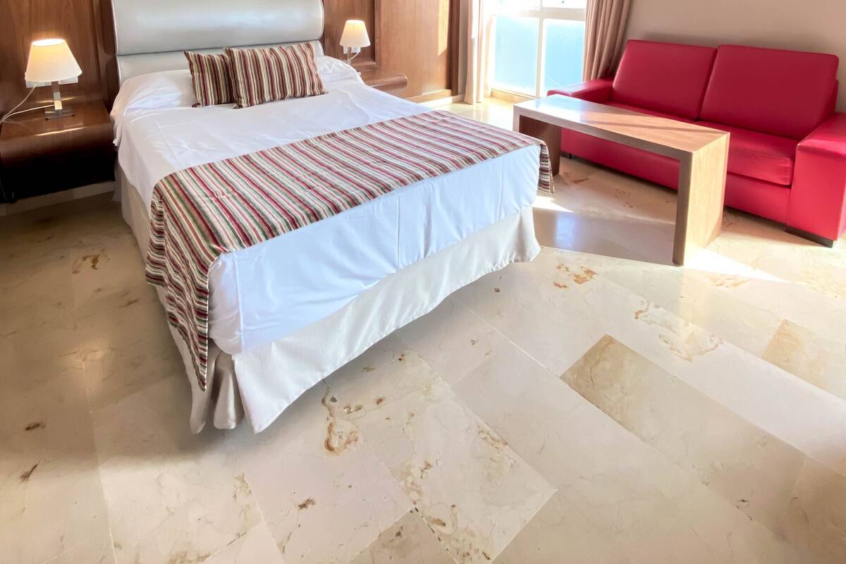 A hotel room with a large bed, a red sofa, a small table, and a window with curtains.