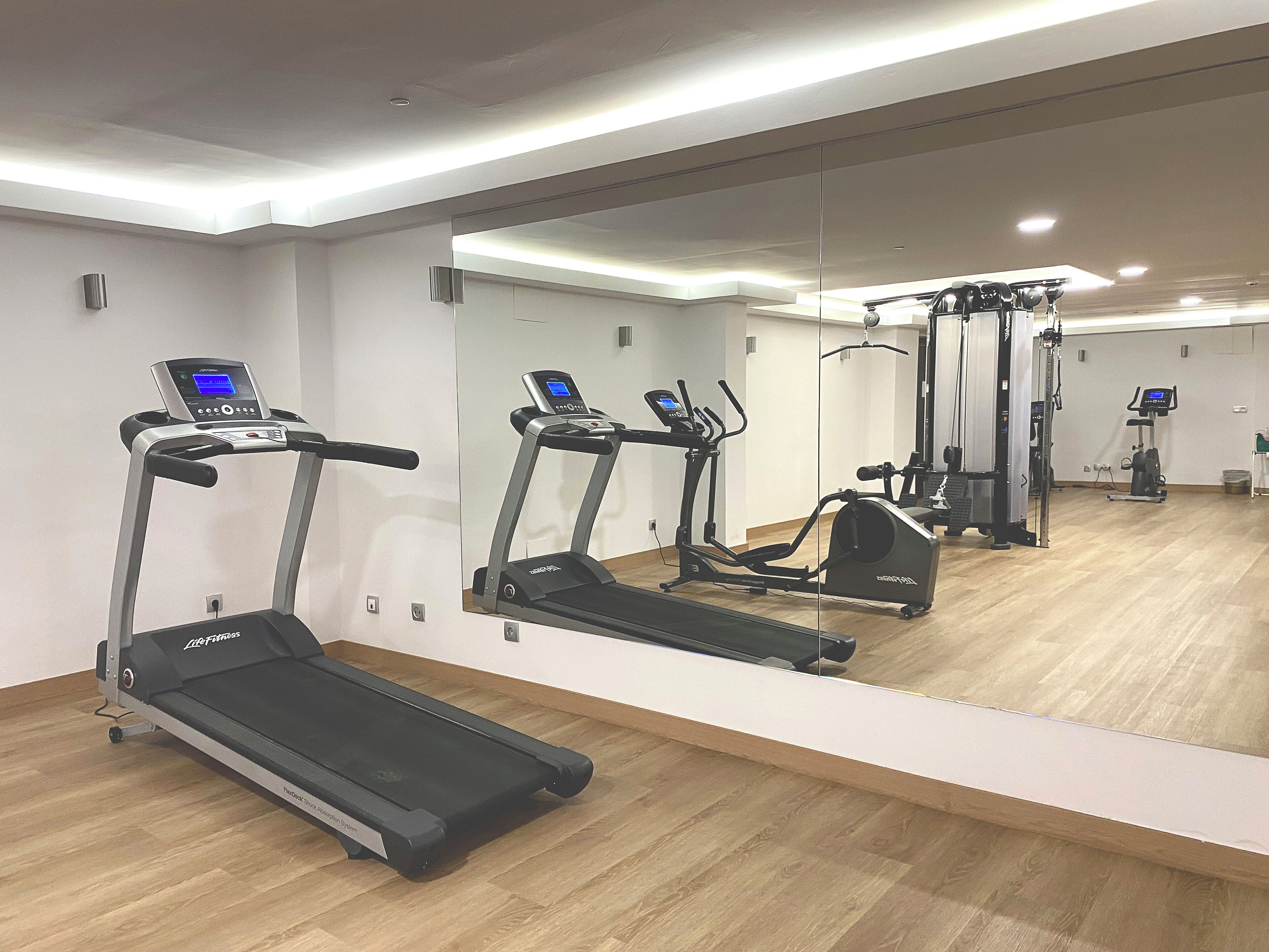 Une salle de sport équipée de tapis de course et d’appareils d’exercice, avec un grand miroir et un sol en bois.