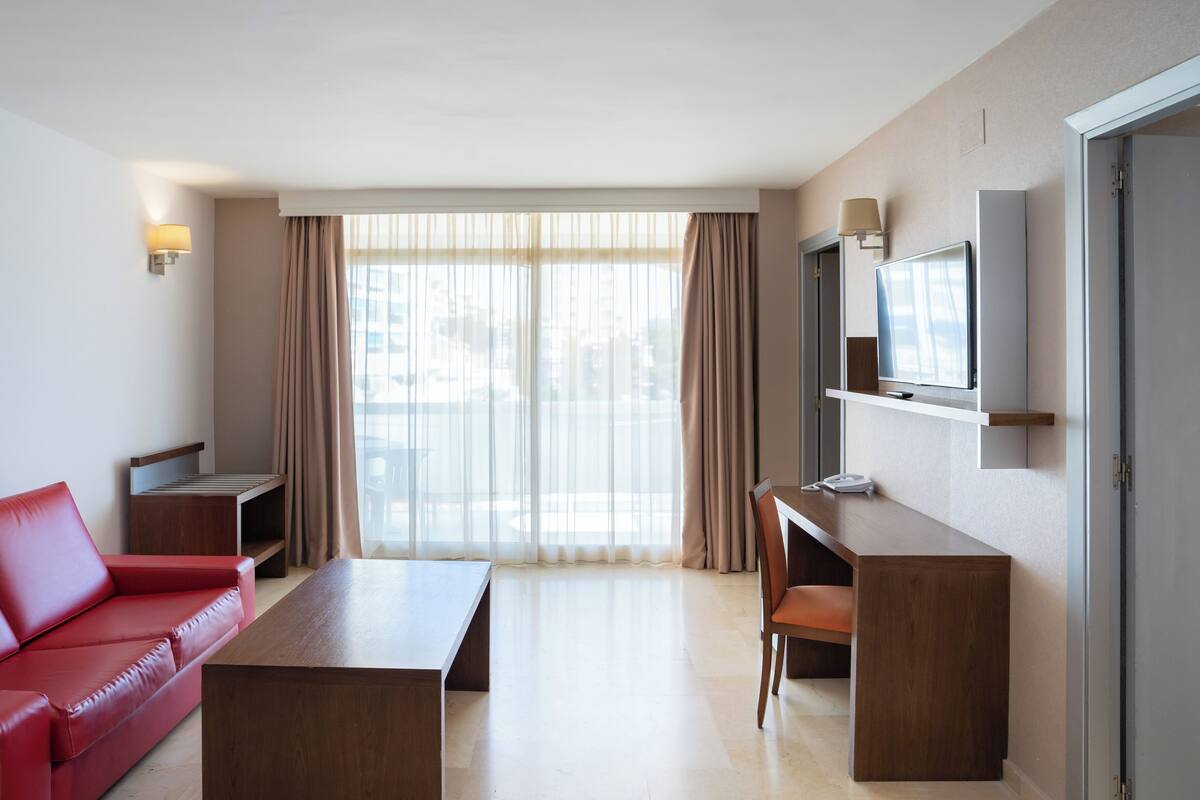 A hotel room with a red sofa, a wooden desk, a television, and a large window with curtains.